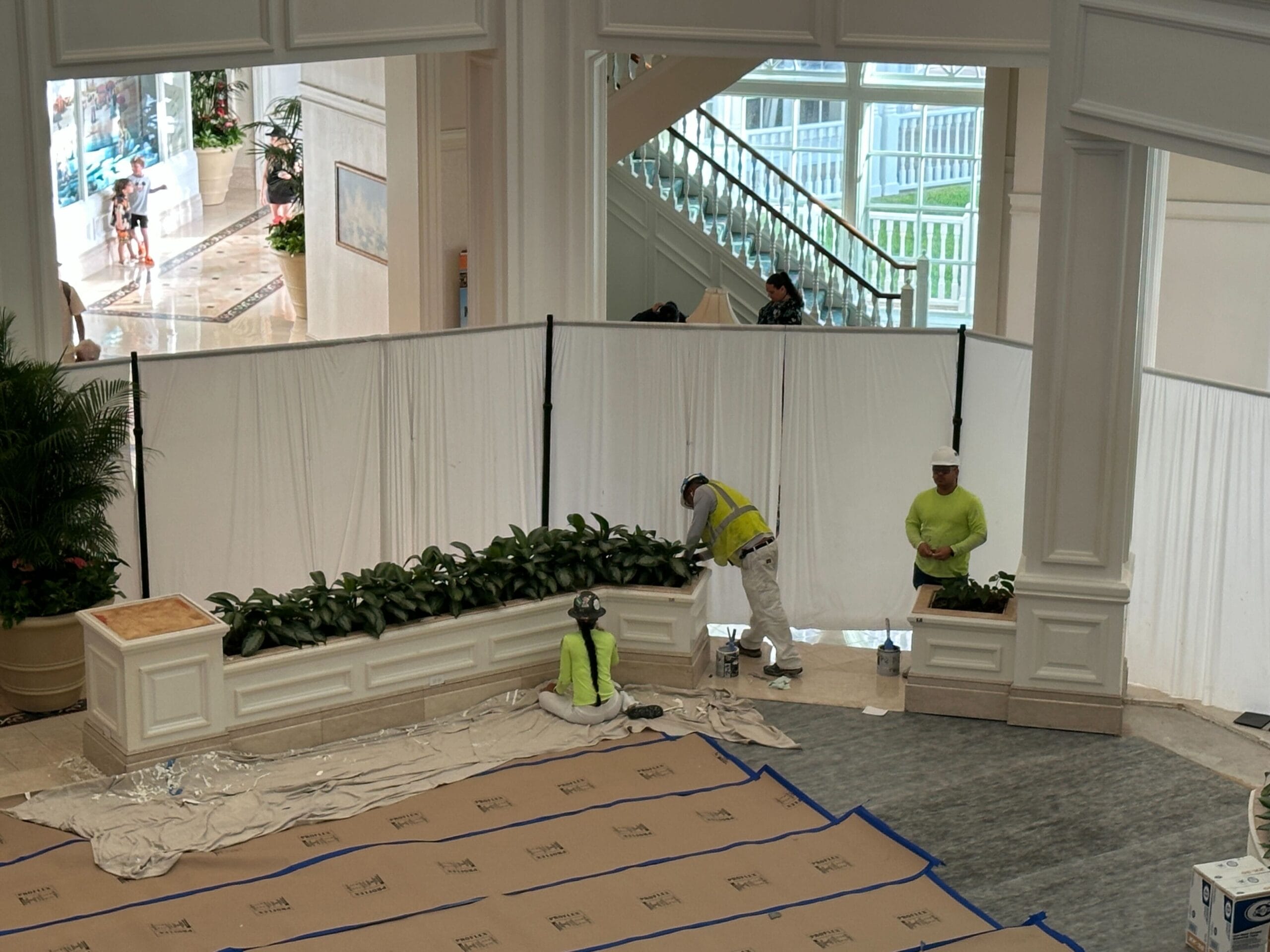 Three workers in safety gear paint near plants behind white screens at Disney’s Grand Floridian Resort, no castle visible.