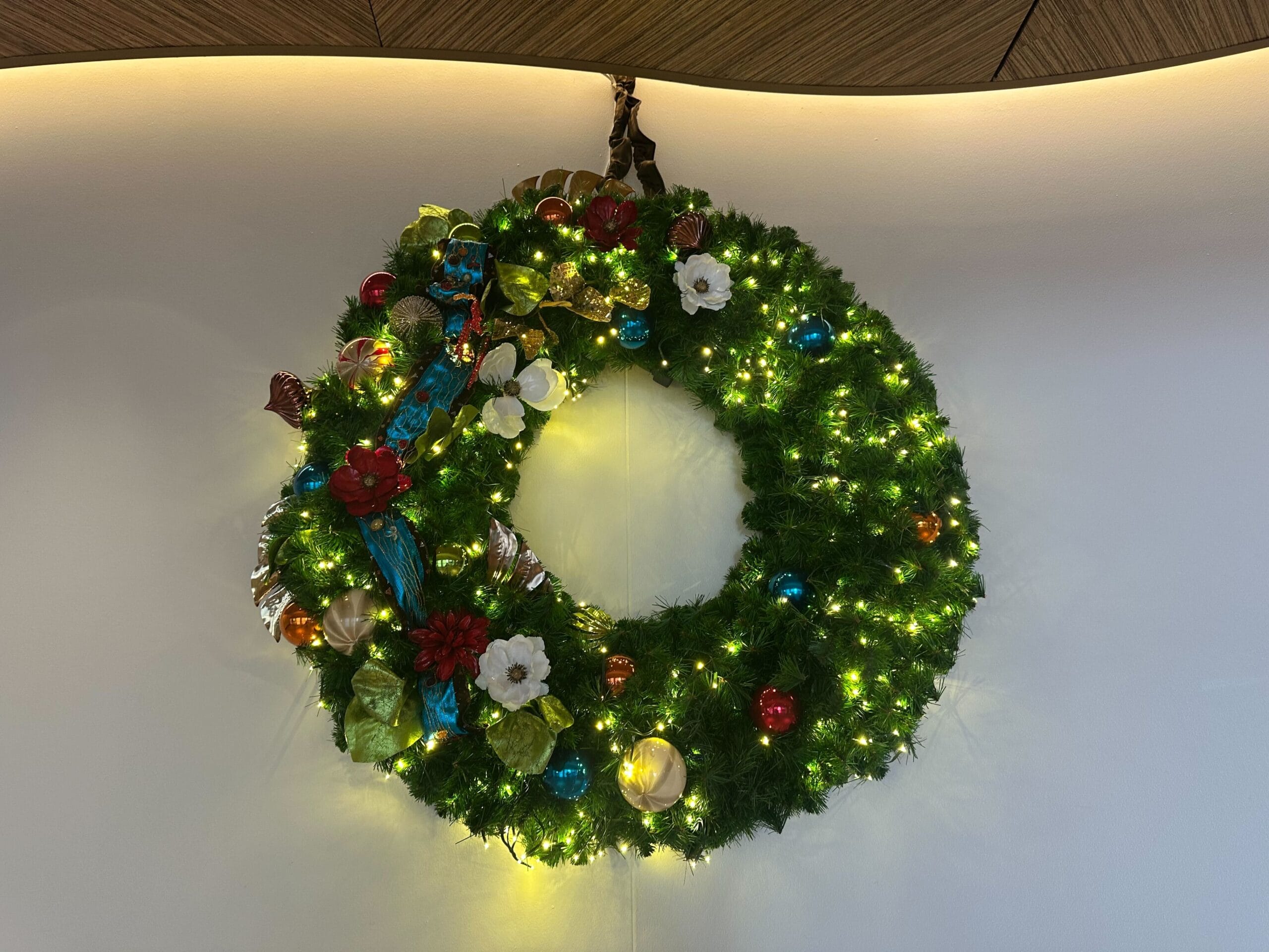 A decorated holiday wreath with green foliage, colorful ornaments, and glowing lights is hanging on a wall.