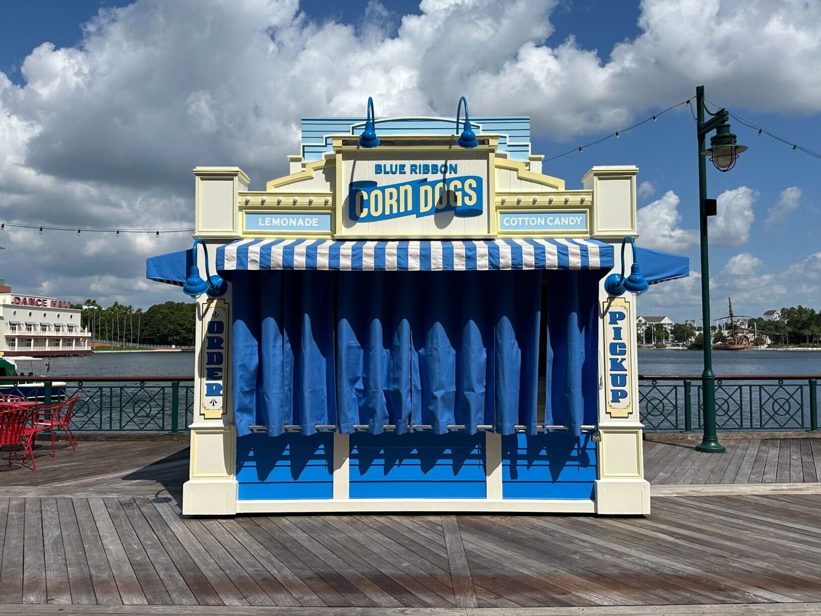 A closed corn dog stand with blue and white striped awnings, labeled Blue Ribbon Corn Dogs, on a boardwalk under a cloudy sky. Red outdoor chairs and tables are visible on the left.