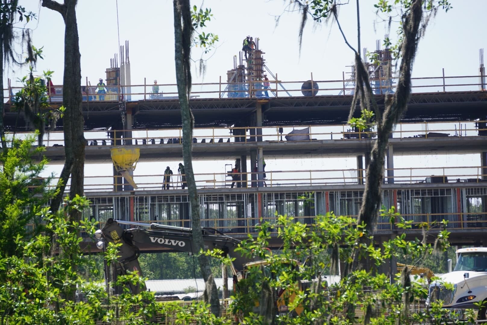 Amidst lush green vegetation, workers on scaffolding are diligently building a multi-story structure at the future Lakeshore Lodge. An excavator is busy in the foreground, reflecting a promising new chapter in this Disney construction update.