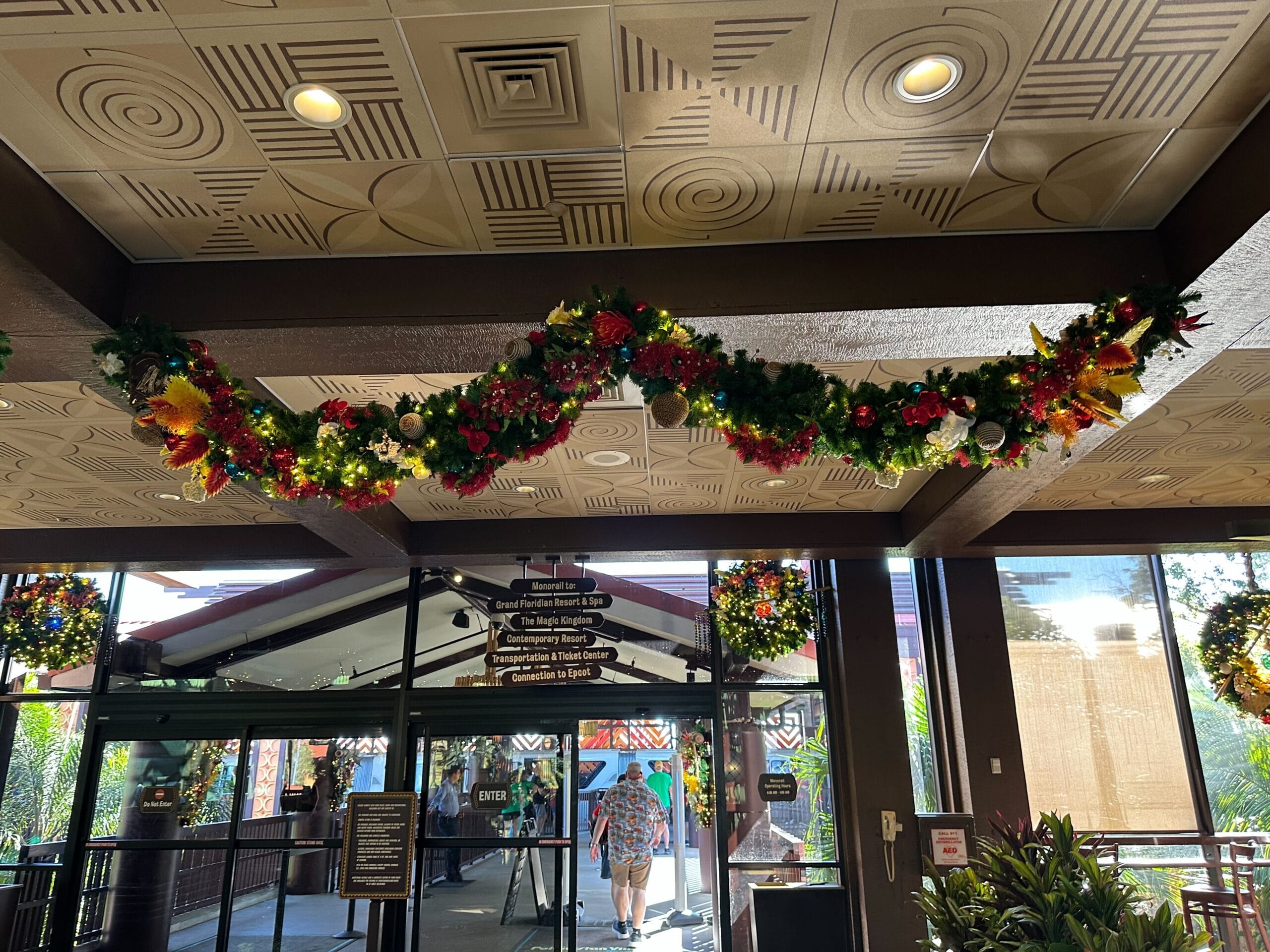 Holiday garland with lights and ornaments hangs from a ceiling. Doors and directional signs are visible below, leading outside.