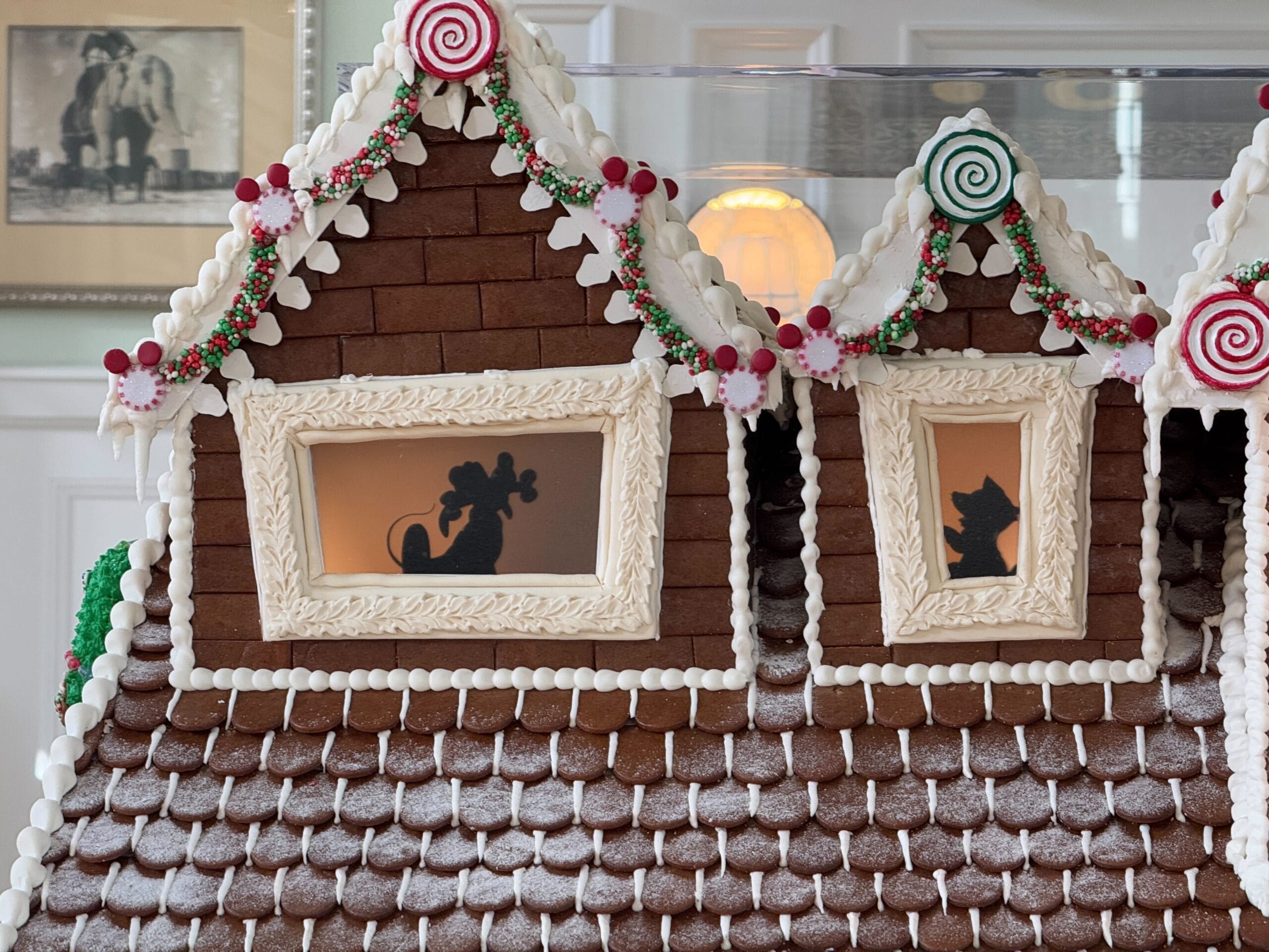 A gingerbread house with two silhouettes in the windows: one of a woman sitting, and the other of a cat. The house is decorated with candy and icing.