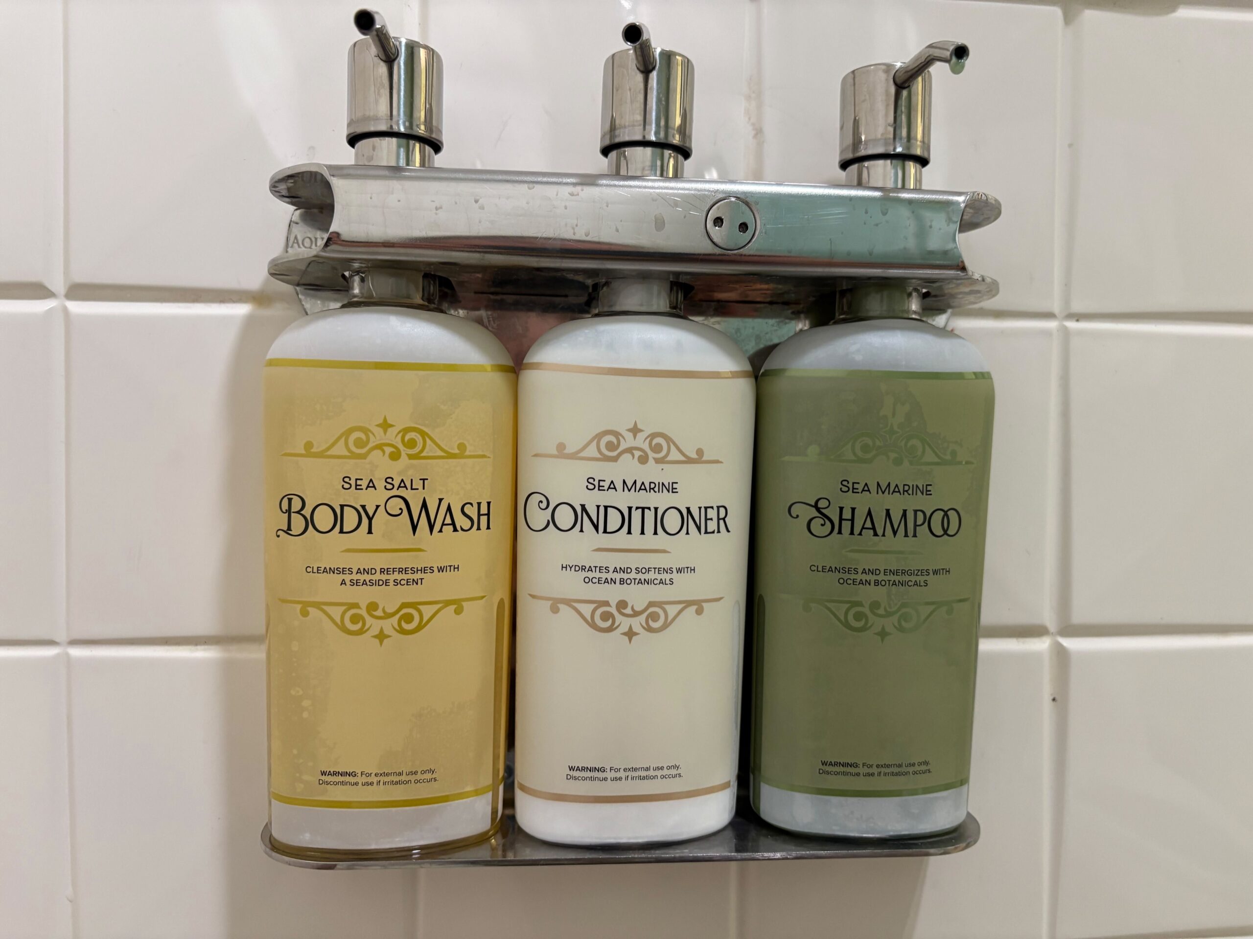 Three wall-mounted dispensers in a bathroom hold sea salt body wash, sea marine conditioner, and sea marine shampoo, labeled clearly for use. The dispensers are stainless steel with white tiles behind them.