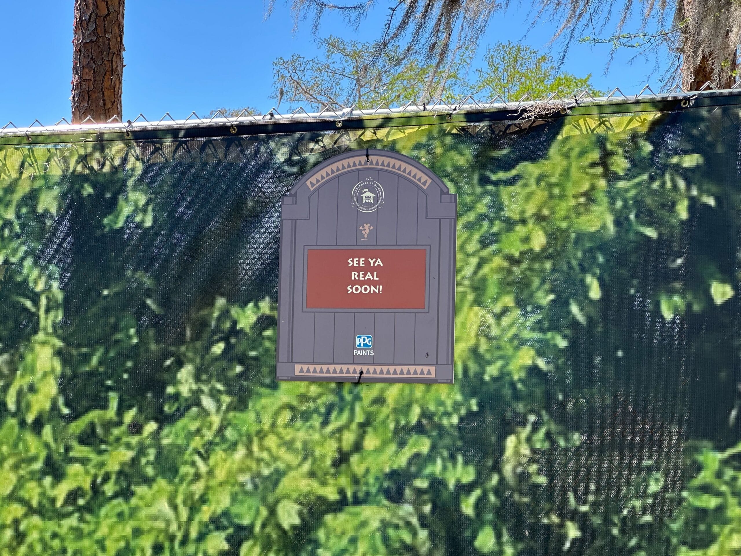 A message on a gate surrounded by greenery reads See ya real soon! with a logo below.