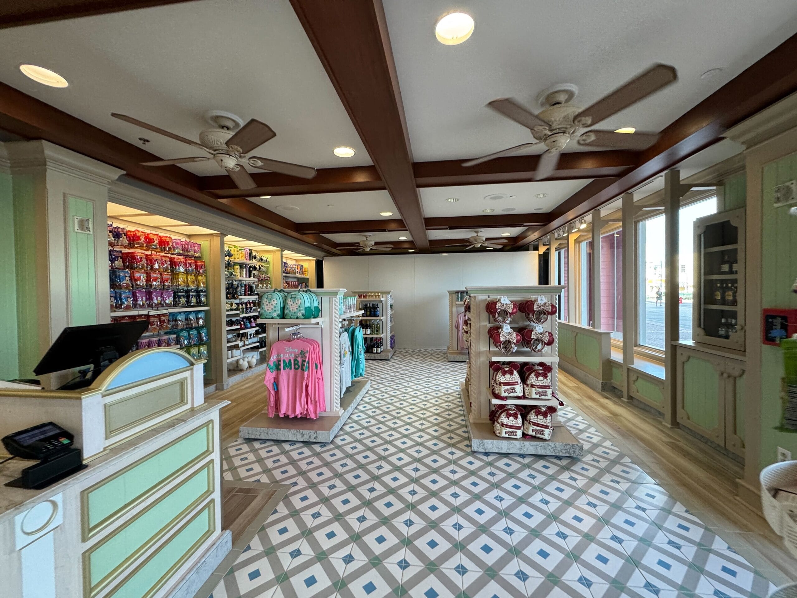 A well-lit store interior with wooden beams, ceiling fans, and patterned floor tiles. Shelves display colorful merchandise including shirts and plush toys. Counter with register on the left.