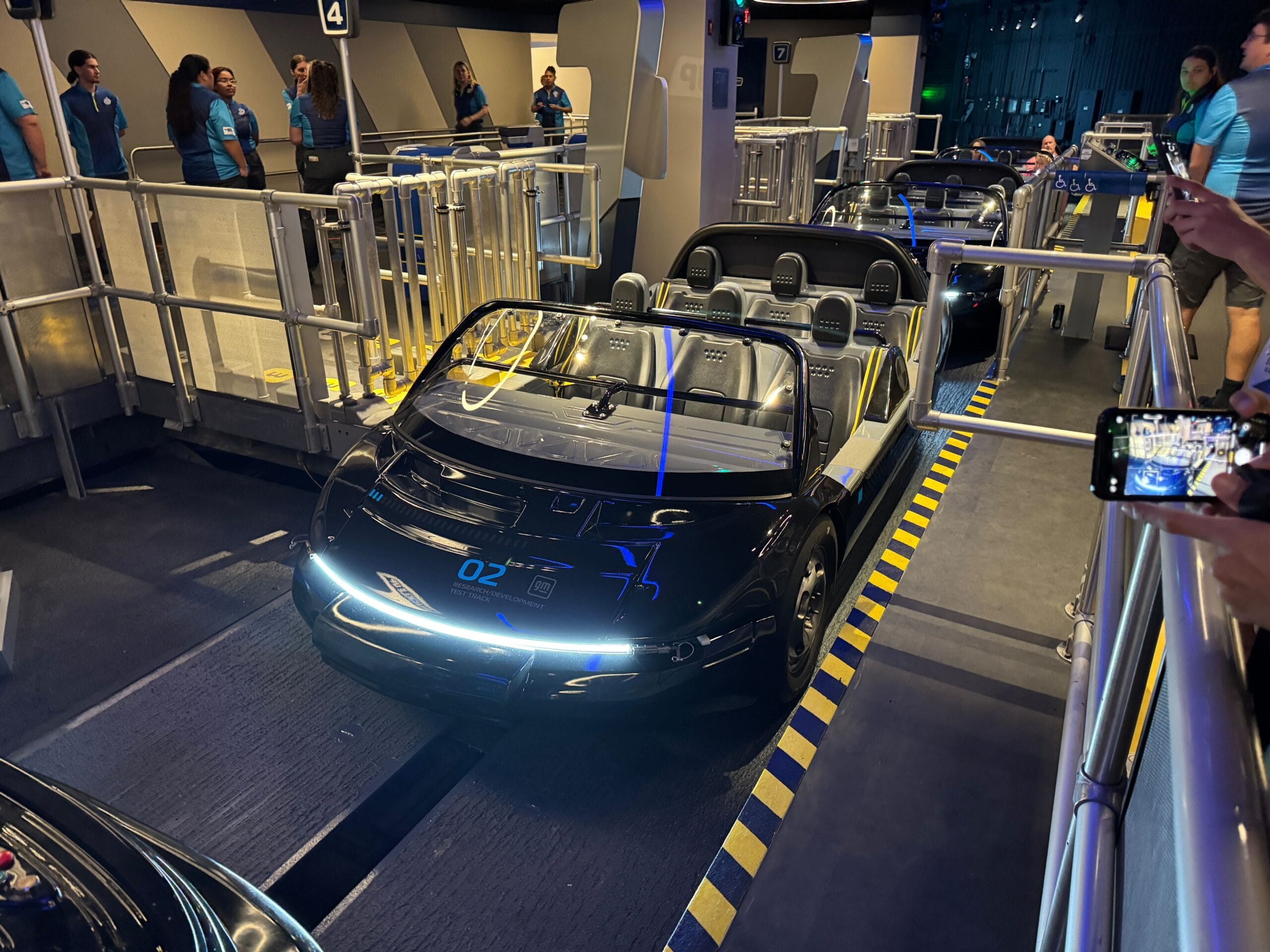 A sleek black Test Track ride vehicle waits at a theme park loading platform with guests and staff nearby.
