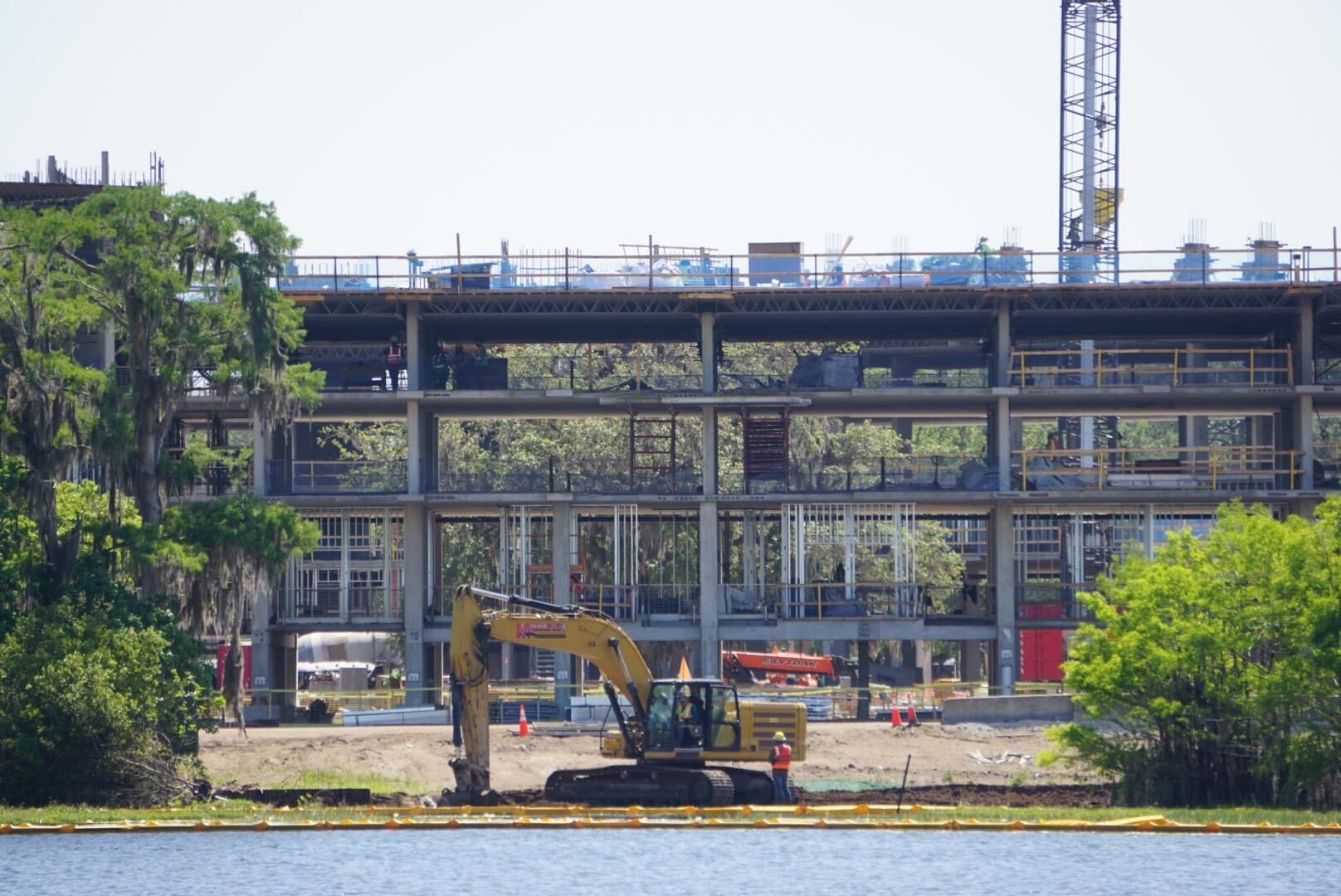 The construction site, near the shimmering Disney Lakeshore Lodge, showcases a prominent building framework with an excavator poised in the foreground. Lush trees flank both sides, hinting at a harmonious blend of nature and development.