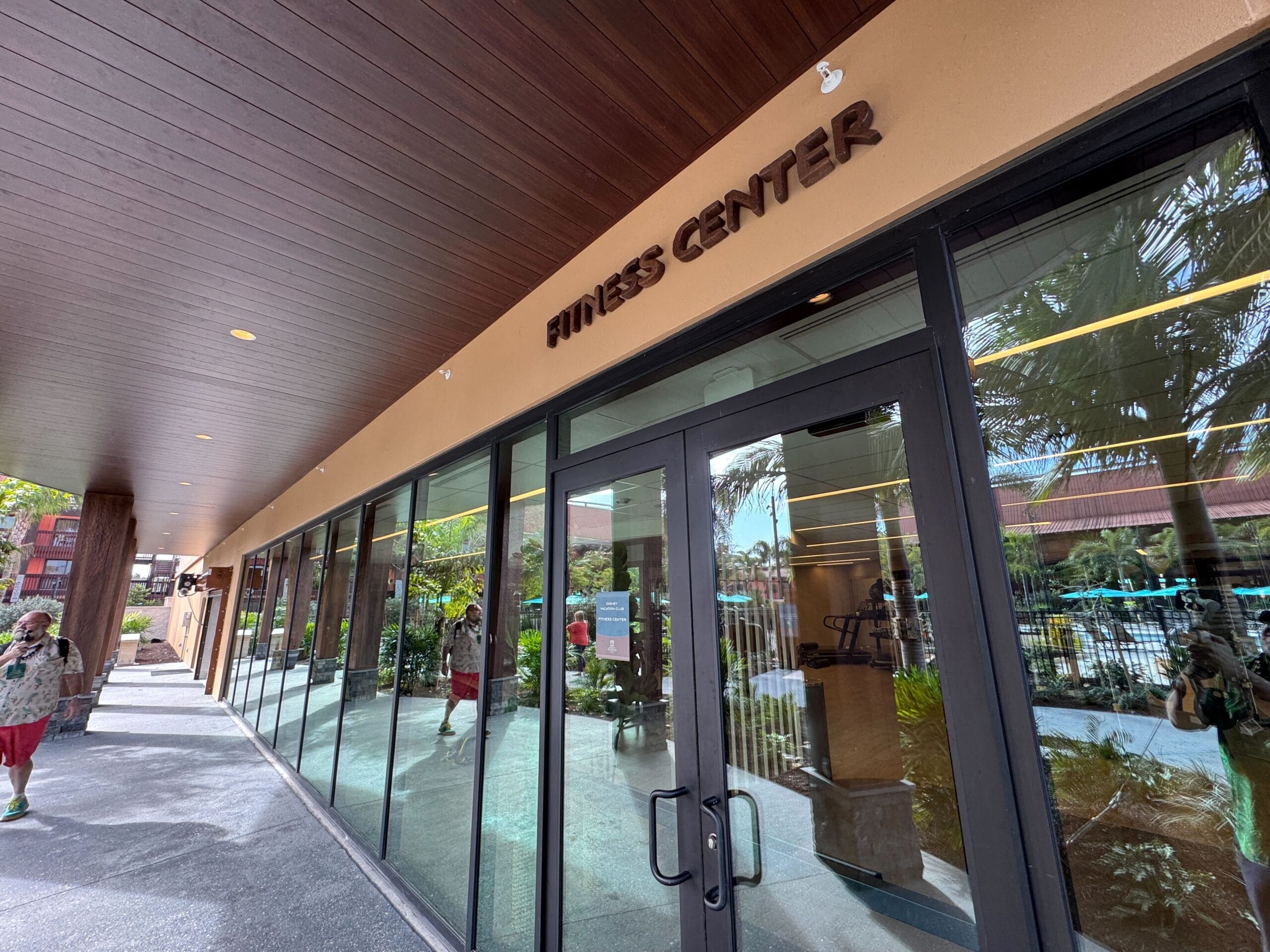 Exterior view of a fitness center with glass doors and windows reflecting a tropical setting.
