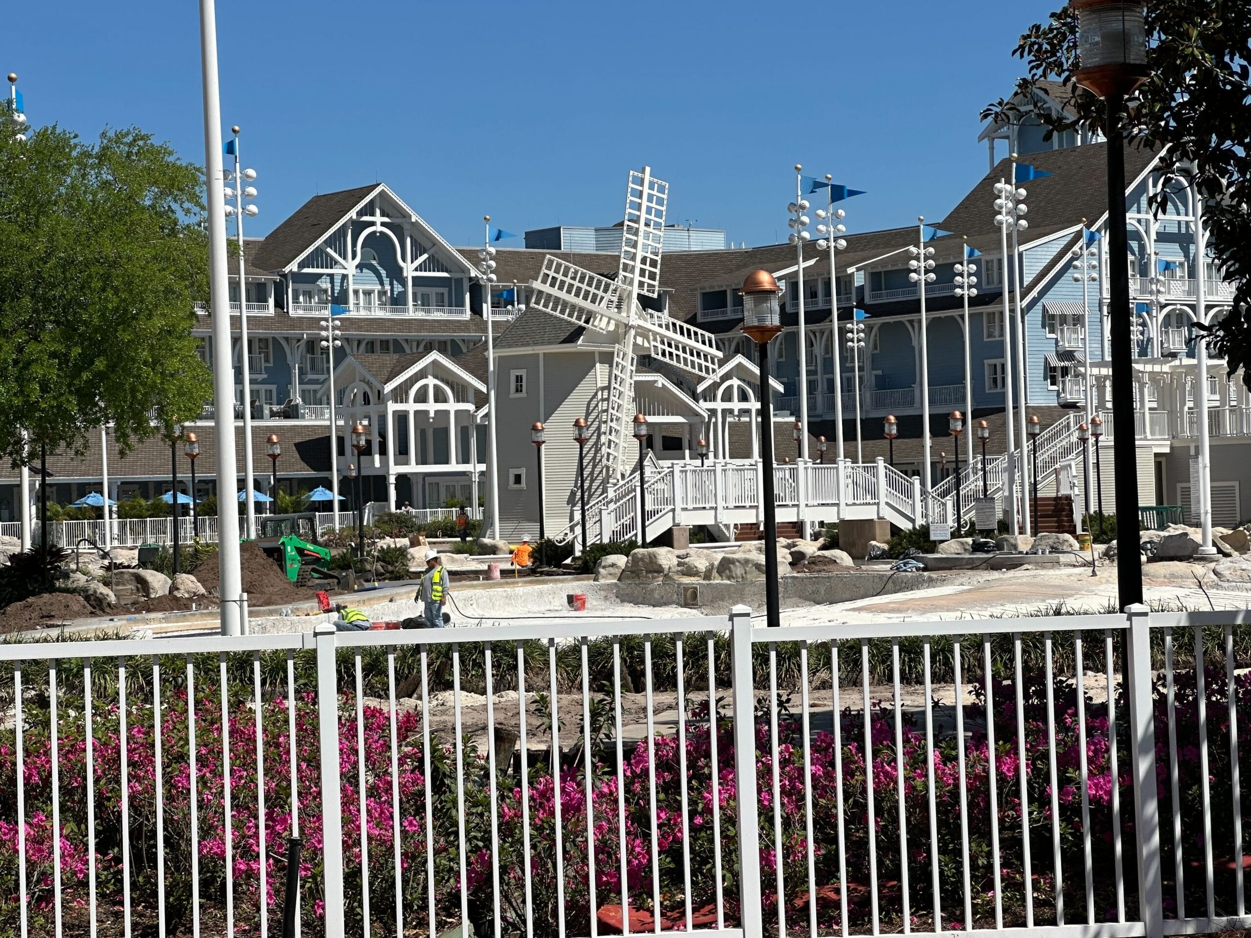 A building with a windmill structure in the foreground, surrounded by a railing, landscaping with flowers, and construction equipment. Clear blue sky in the background.