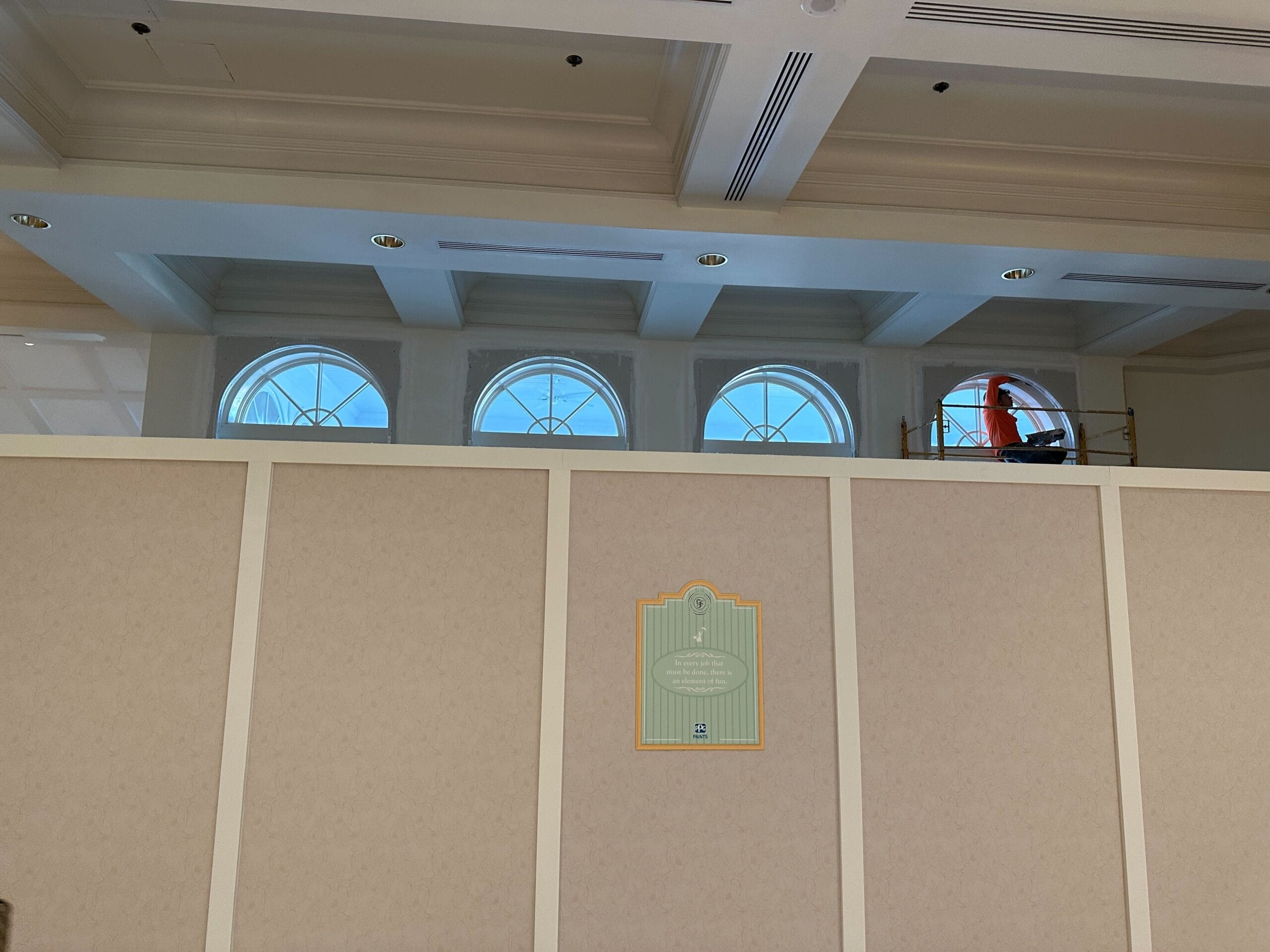 A worker on scaffolding cleans arched windows above a partition in a well-lit room with decorative ceiling beams.