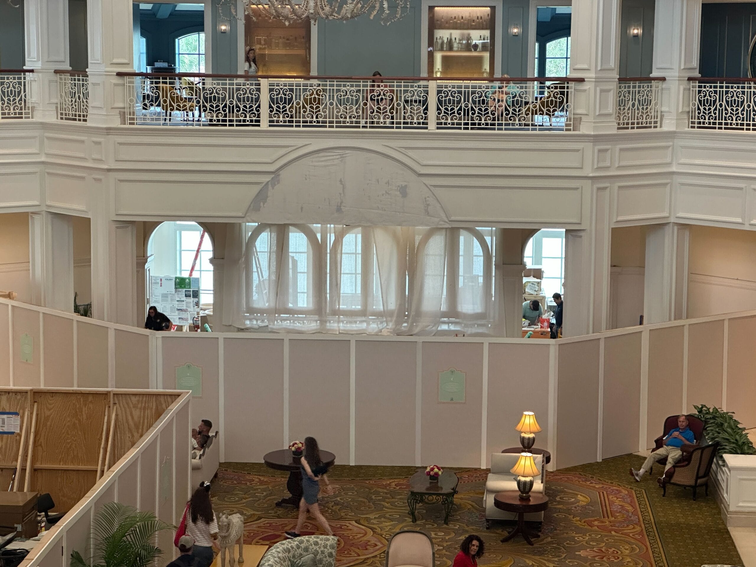 A hotel lobby with construction walls, lounge chairs, people walking, and a second-floor balcony above.