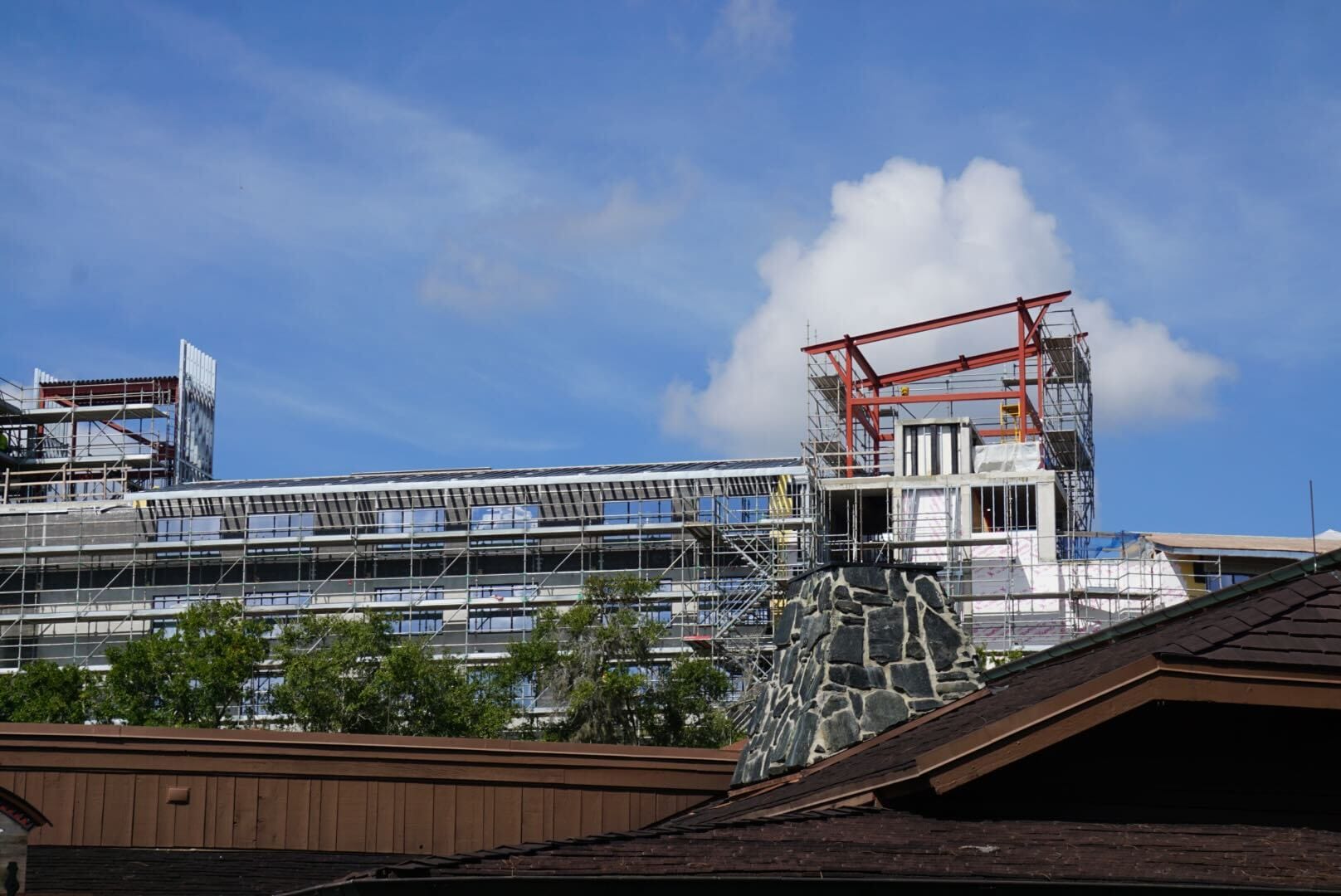 A building under construction with scaffolding at Disney Lakeshore Lodge, peeking above rooftops and trees under a blue sky.