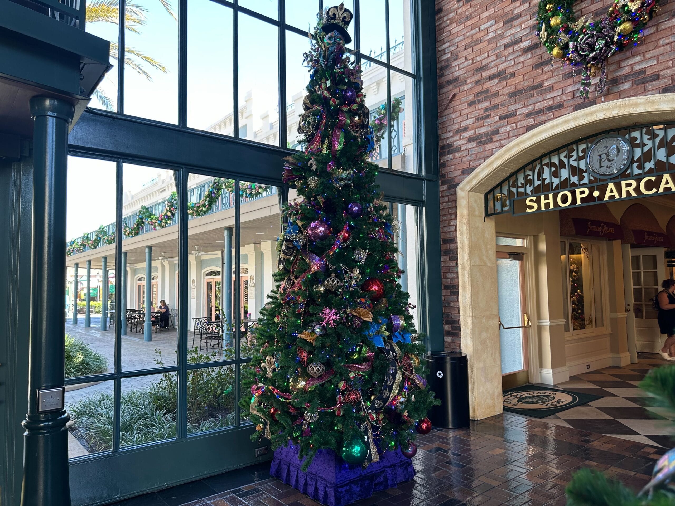 Holiday-themed decorated Christmas tree in a shopping arcade, adorned with ornaments and ribbons, stands by large windows overlooking an outdoor area.