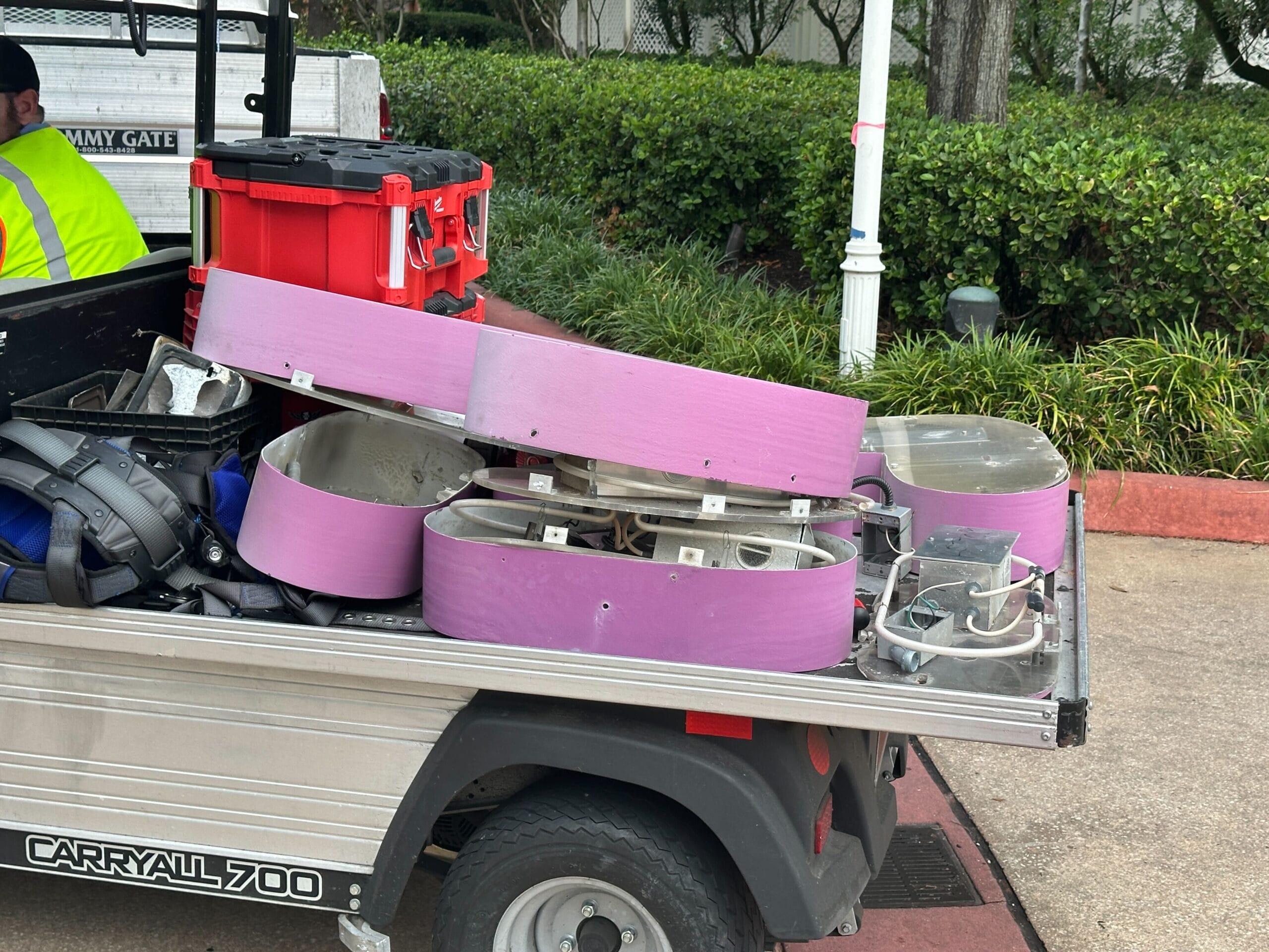 Several purple and clear plastic light fixtures are piled in the back of a small utility vehicle near some bushes and a sidewalk.