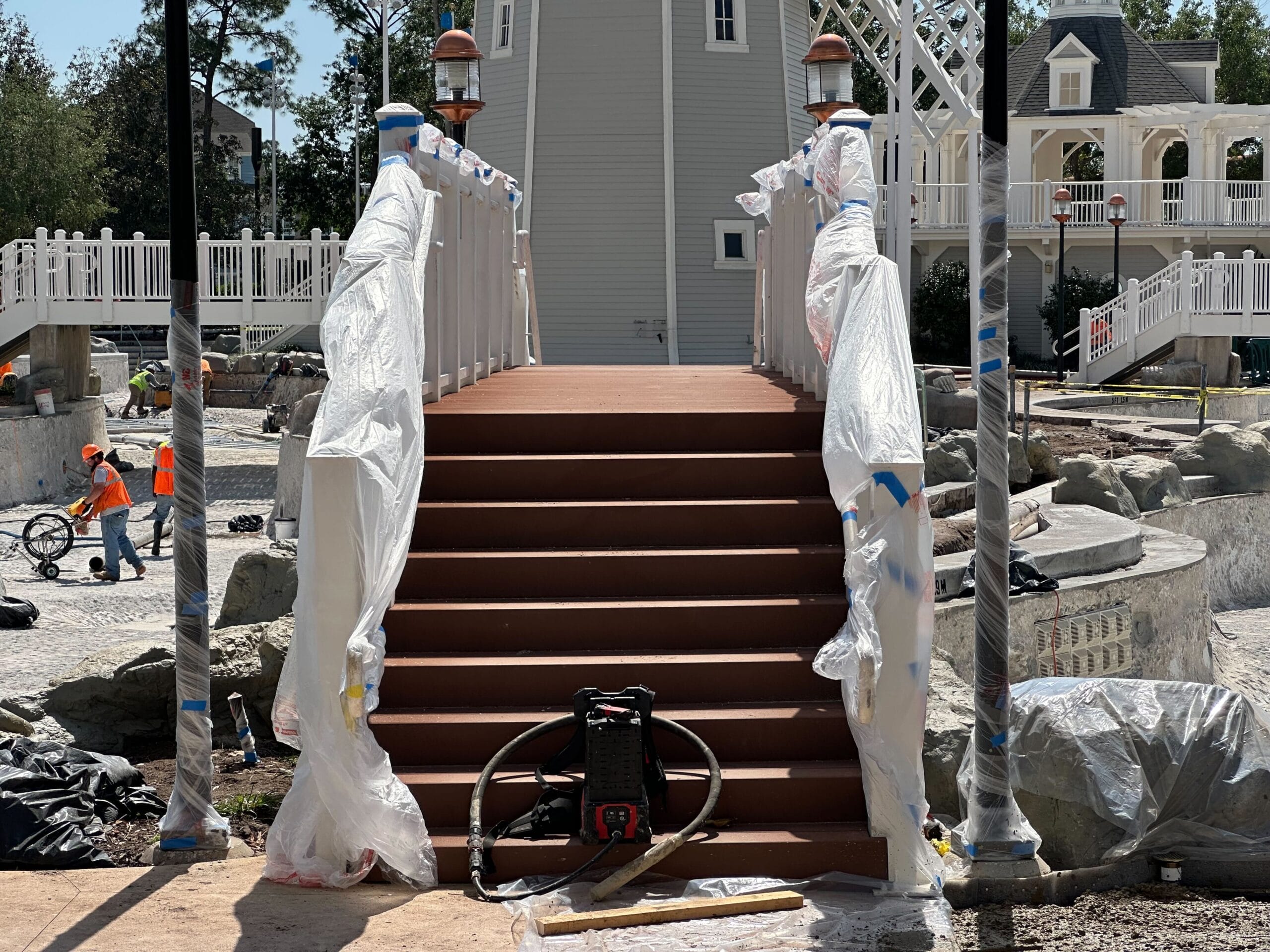 A small brown bridge under construction, wrapped in protective plastic and surrounded by scaffolding. Workers in orange vests are around, with tools and equipment visible. In the background, a gray building and white fences suggest proximity to Disney’s Yacht Club.