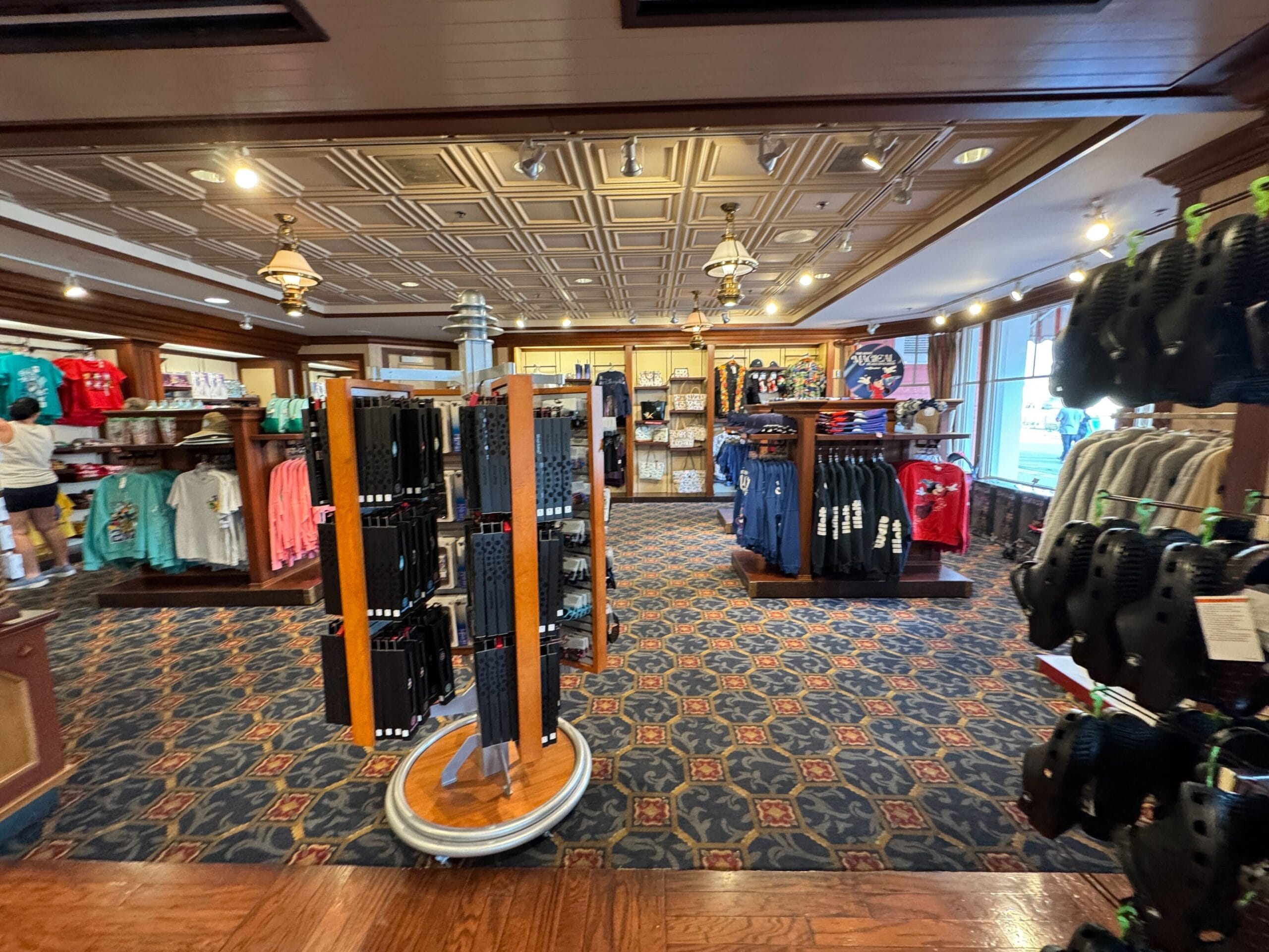 A store interior with racks of clothing, including t-shirts and sweaters, and a display of black shoes. The room has patterned carpet and a decorative ceiling. Lighting appears warm and inviting.