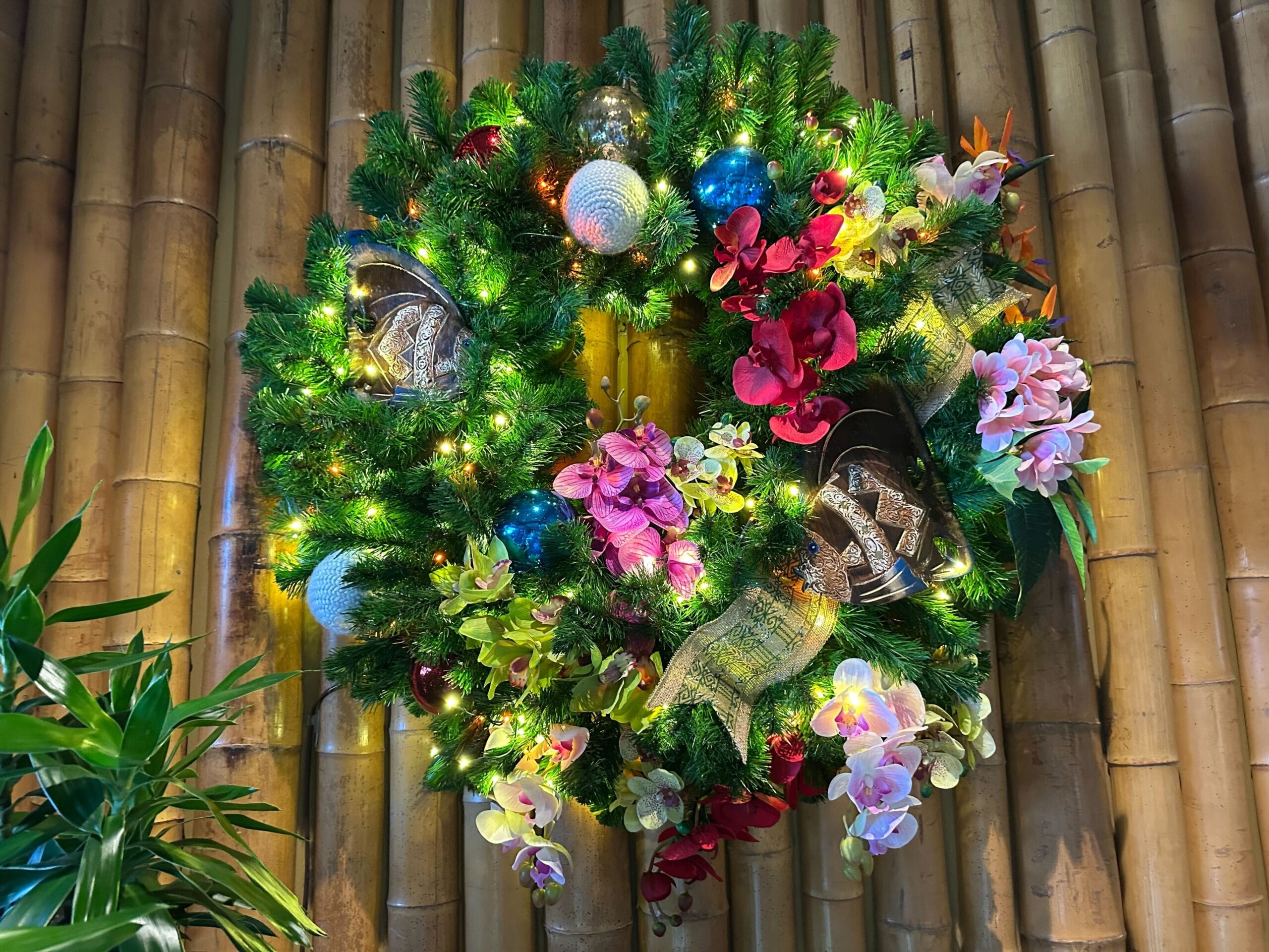 A vibrant, illuminated wreath with red, pink, and white flowers, assorted ornaments, and greenery, hangs on a bamboo wall.