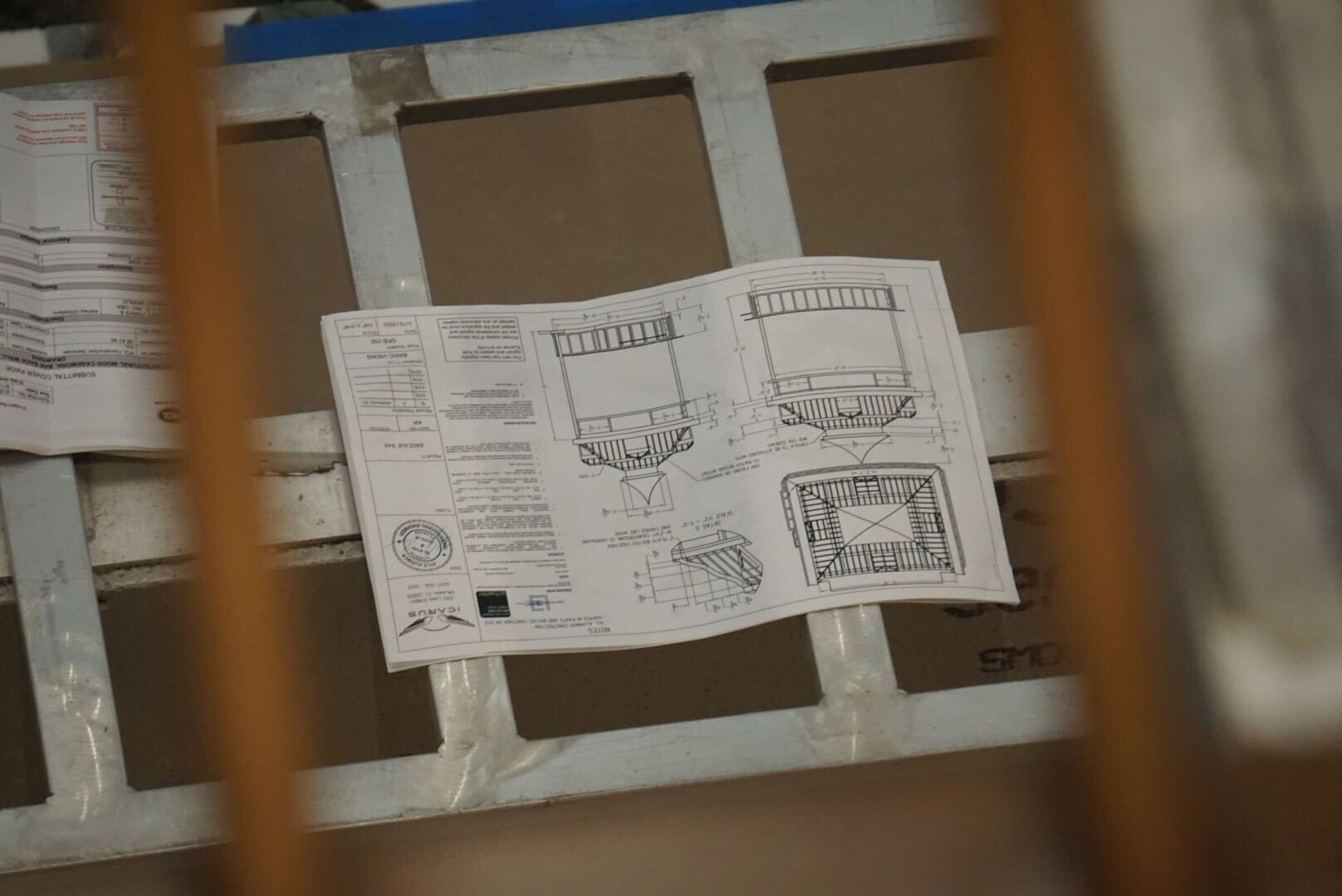 A technical drawing with diagrams and notes rests on a metal grid surface, viewed through vertical bars.