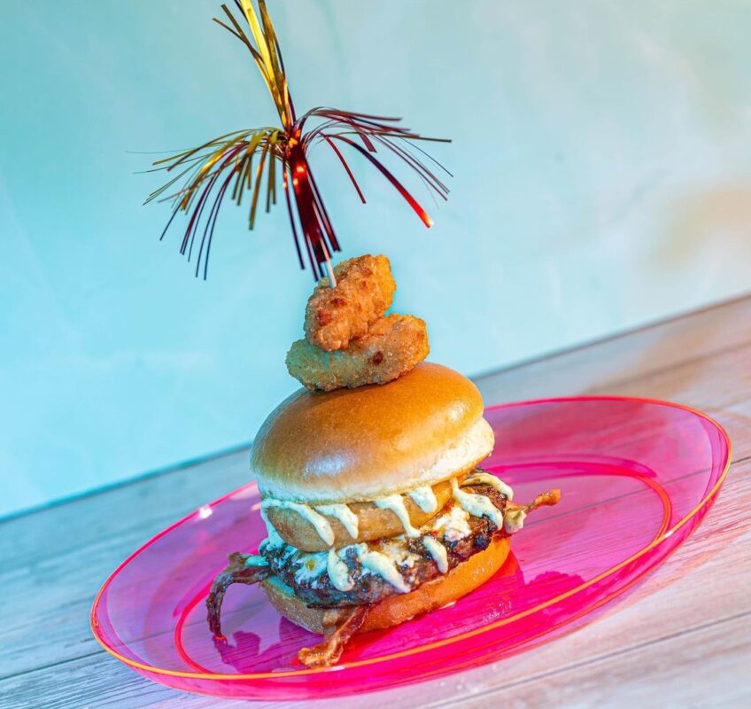 A decorated hamburger featuring a bun, patty, onion ring, cheese, and two chicken nuggets on top, garnished with a sparkler and served on a pink plate at Disney H2O Glow at Typhoon Lagoon Water Park.