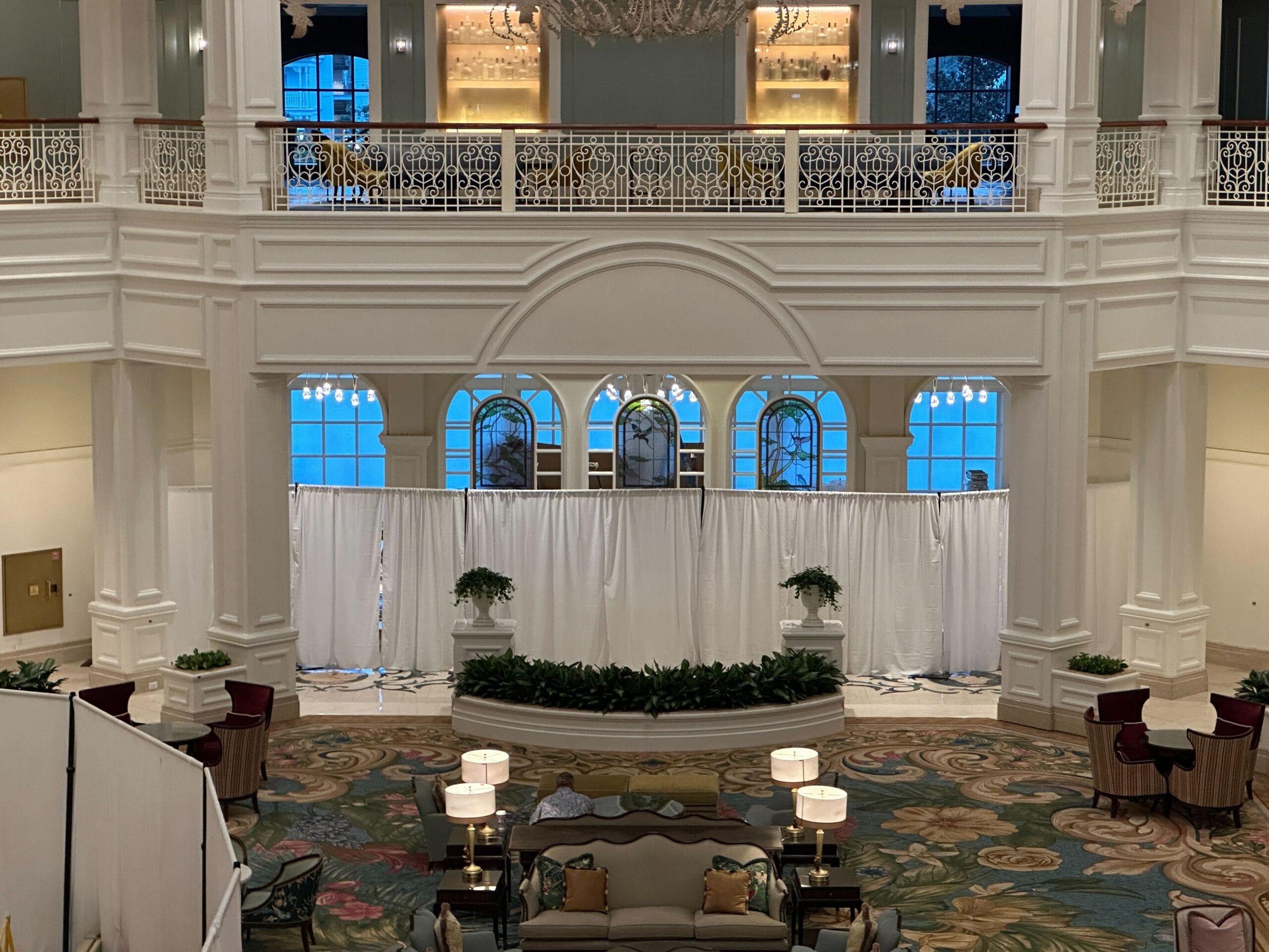 Elegant hotel lobby reminiscent of Disney resort grandeur, with lofty ceilings, chandeliers, and tea room windows by white drapes.