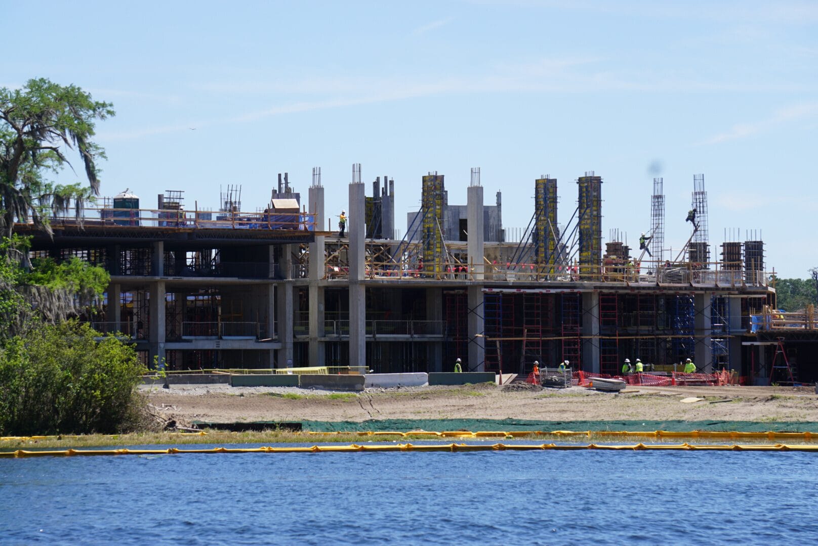 Construction site with partially built multi-story structure, scaffolding, and workers. A body of water is in the foreground.