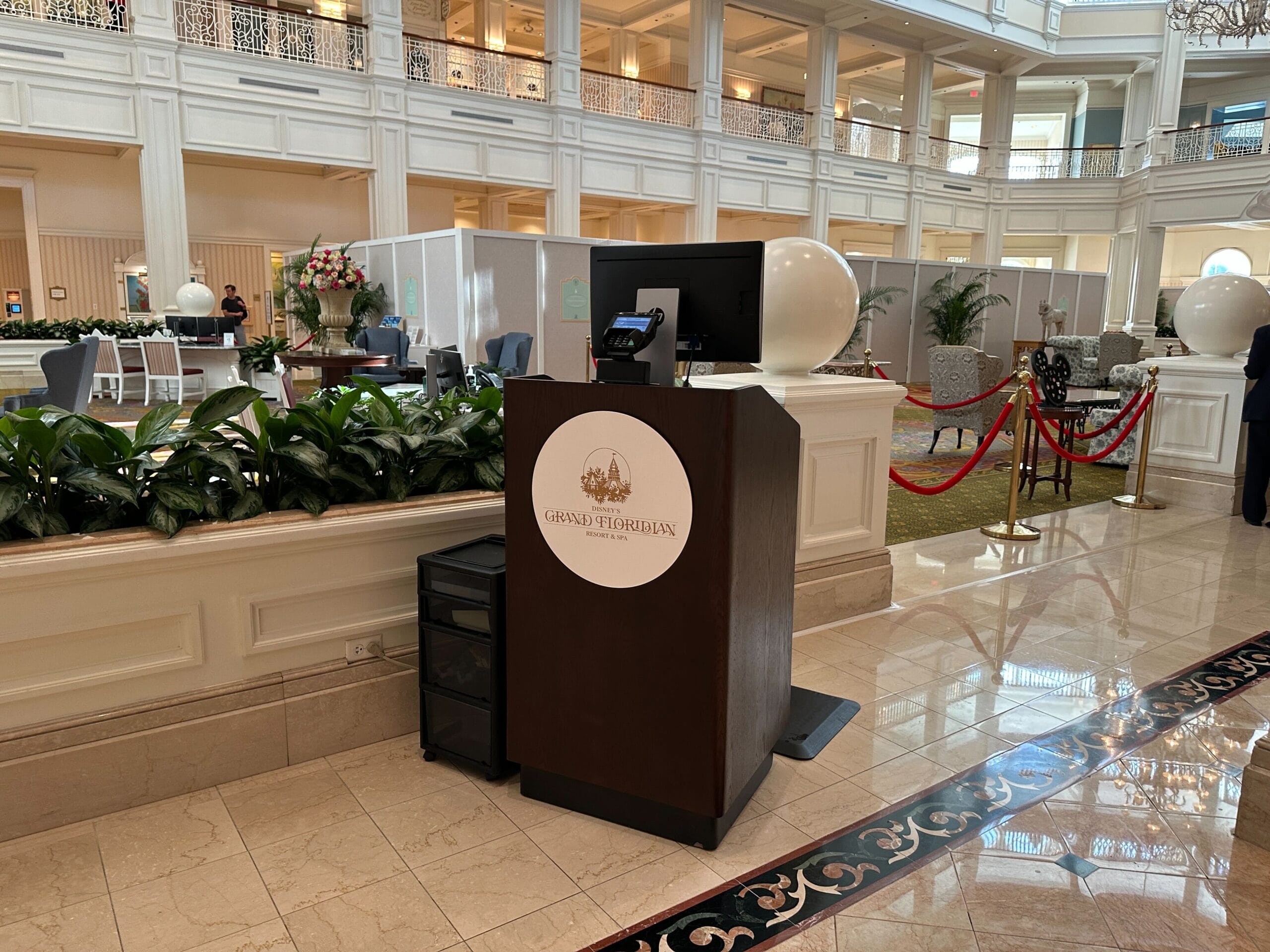 Reception podium in a grand hotel lobby with elegant decor, plants, and a rope barrier in the background.