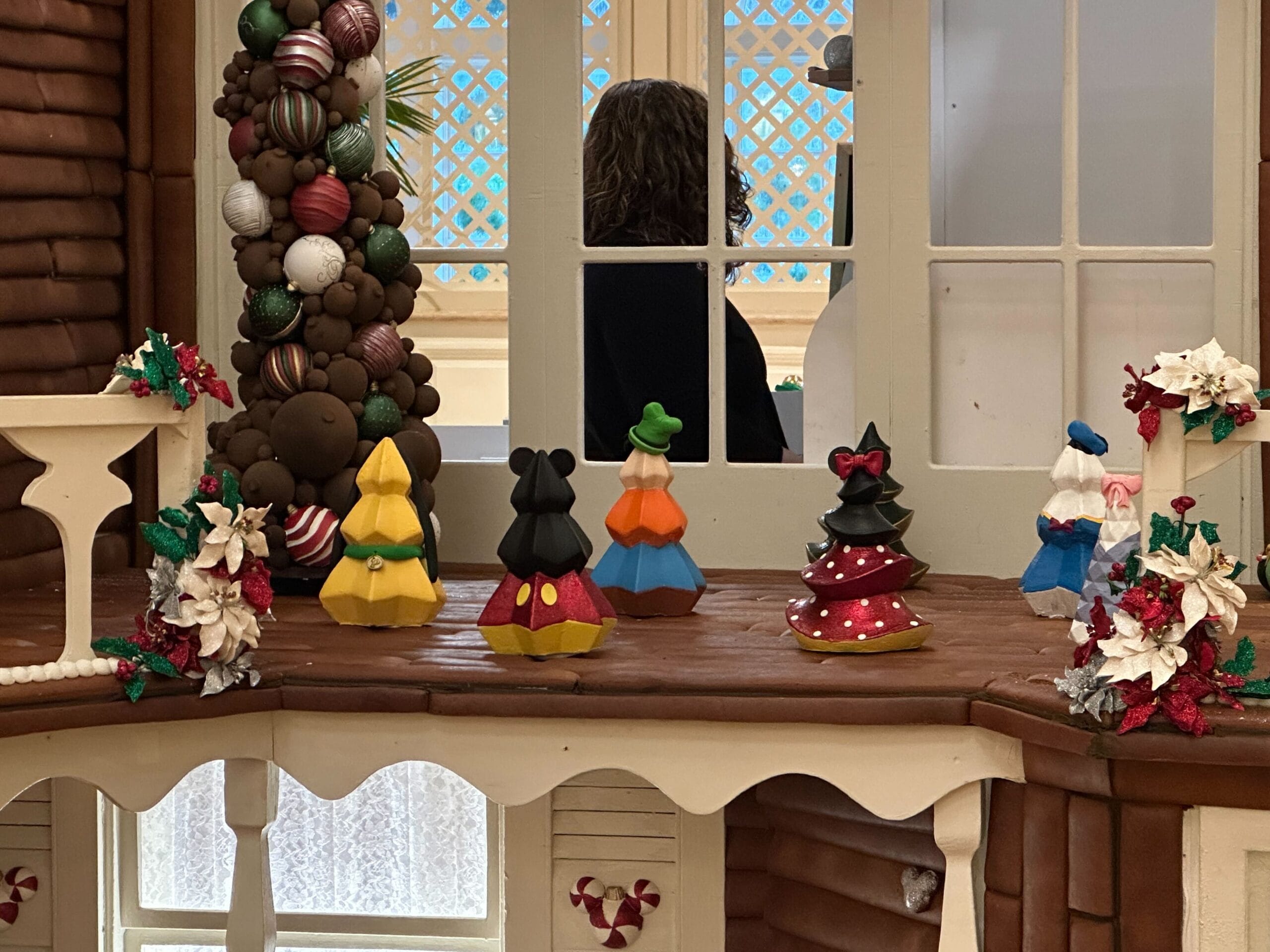 Colorful cone-shaped decorations and floral arrangements adorn a gingerbread house display, with a person visible in the background through a window.