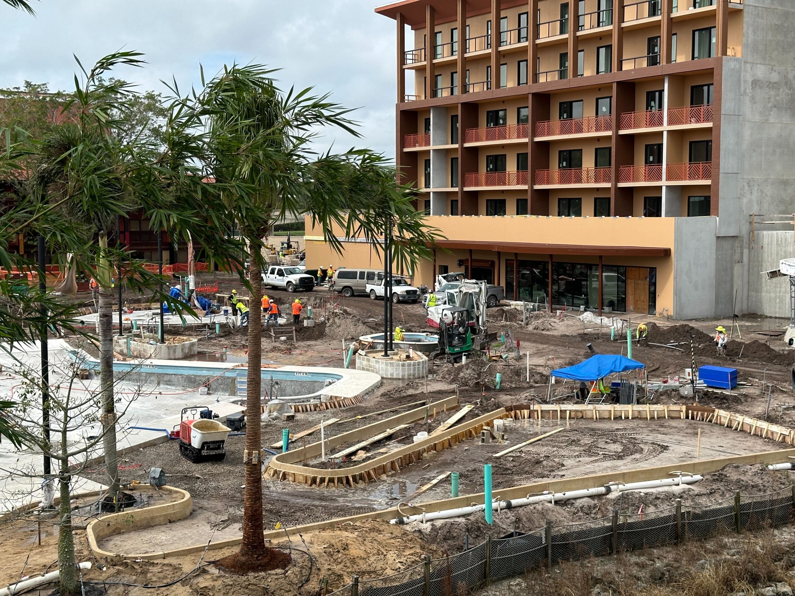 Construction site of a resort featuring an uncompleted pool area, surrounded by workers and equipment. A multi-story building in the background, and a green tree in the foreground.
