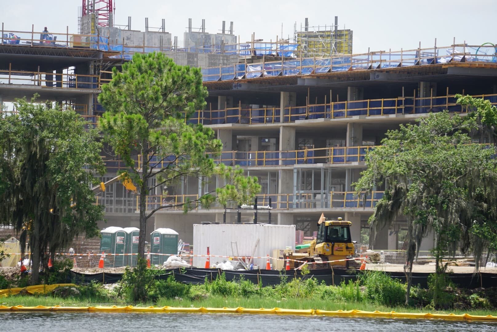 Disney Lakeshore Lodge is under construction near a lake, with trees, equipment, and portable toilets in front.