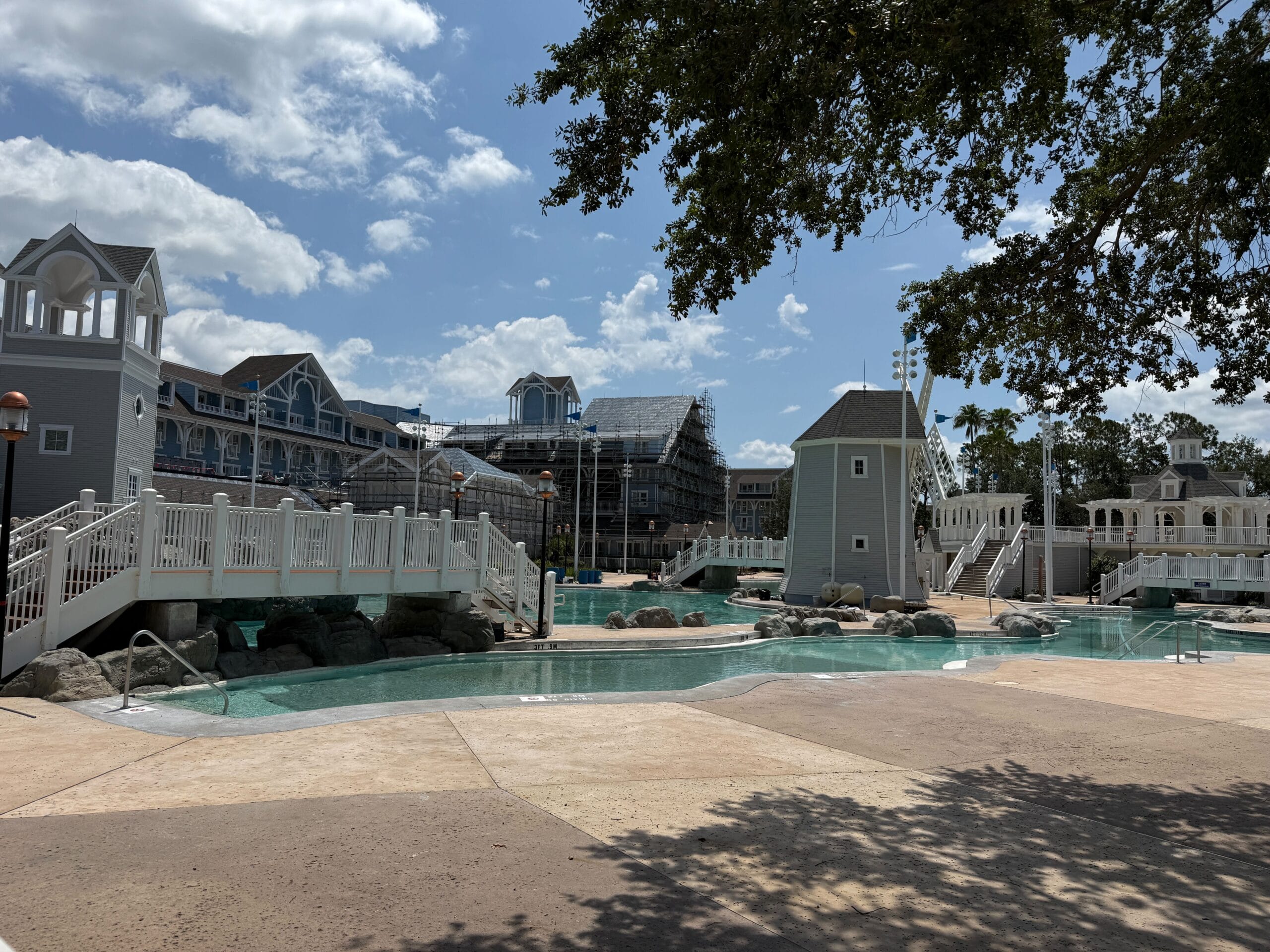A resort pool area with bridges, rocks, and buildings in the background under a partly cloudy sky.