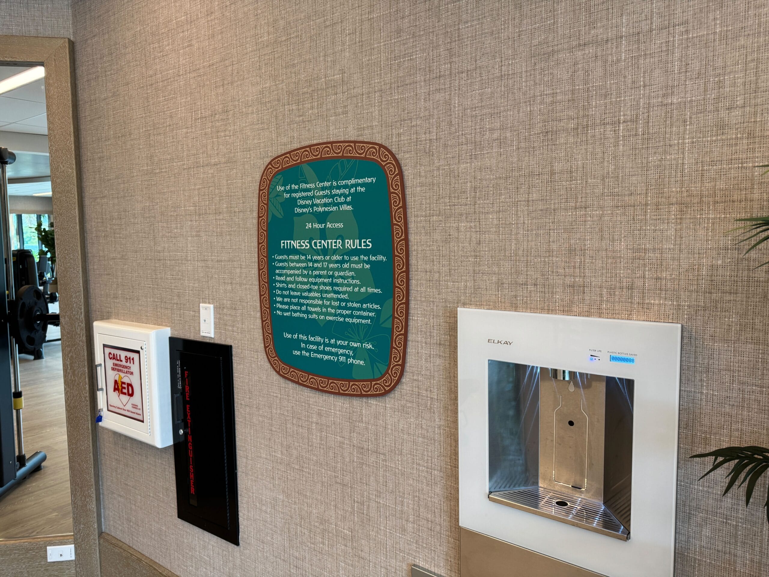 A fitness center wall displaying rules beside a water dispenser and an AED station. The room visible through the doorway has gym equipment.