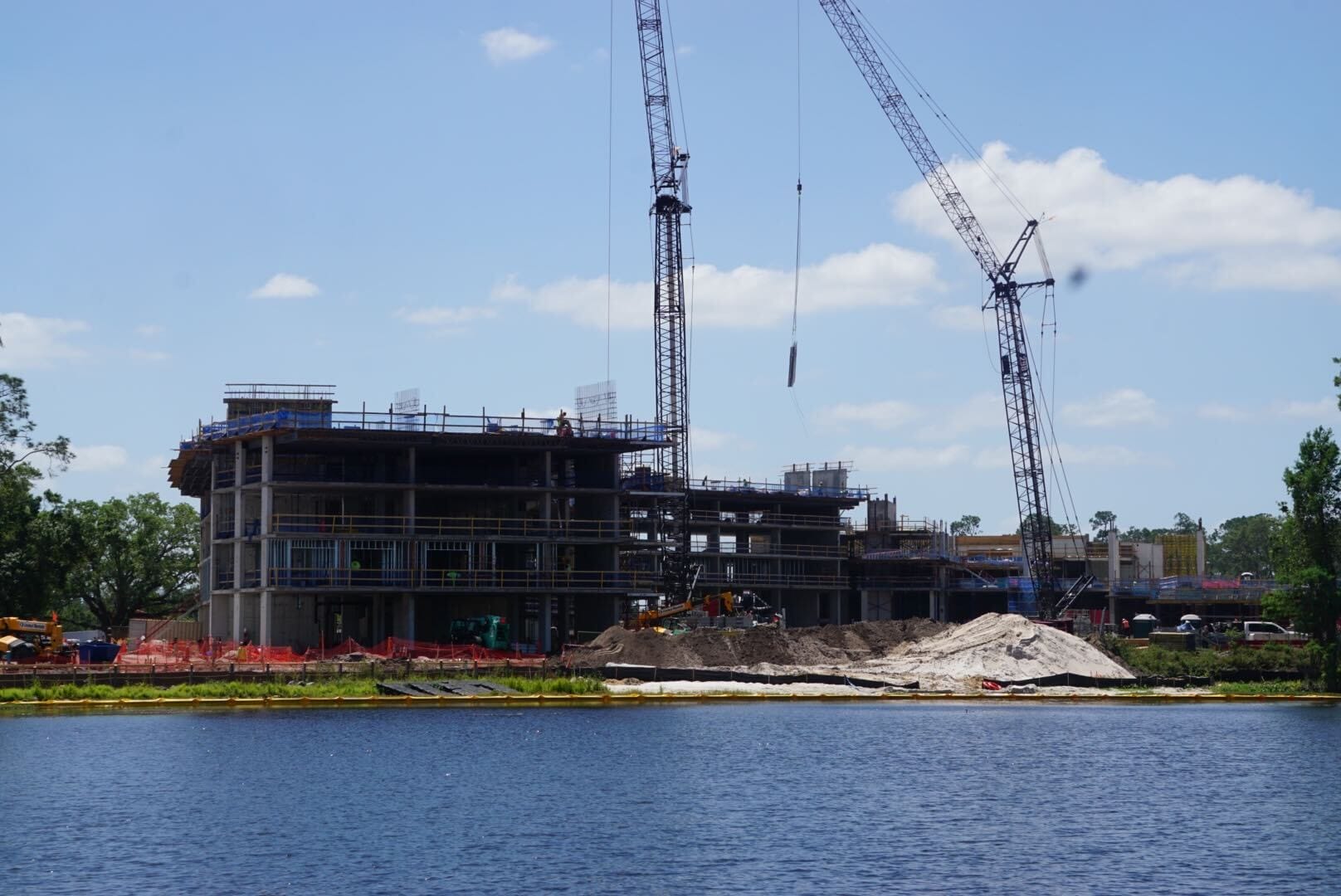 Construction site with a building in progress, two cranes, and piles of sand near a body of water. Trees and a clear blue sky in the background.