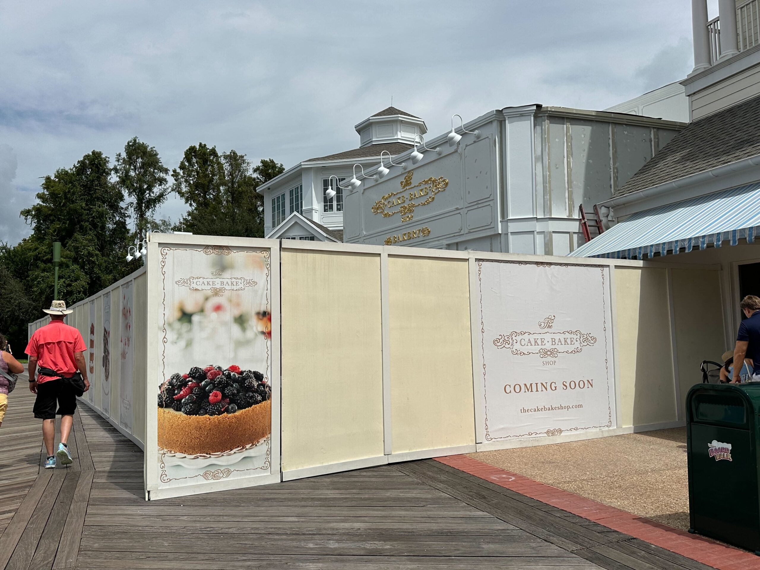 Construction site enclosed by white panels with signs for The Cake Bake Shop that say Coming Soon. The area is located along a boardwalk with some people walking by.