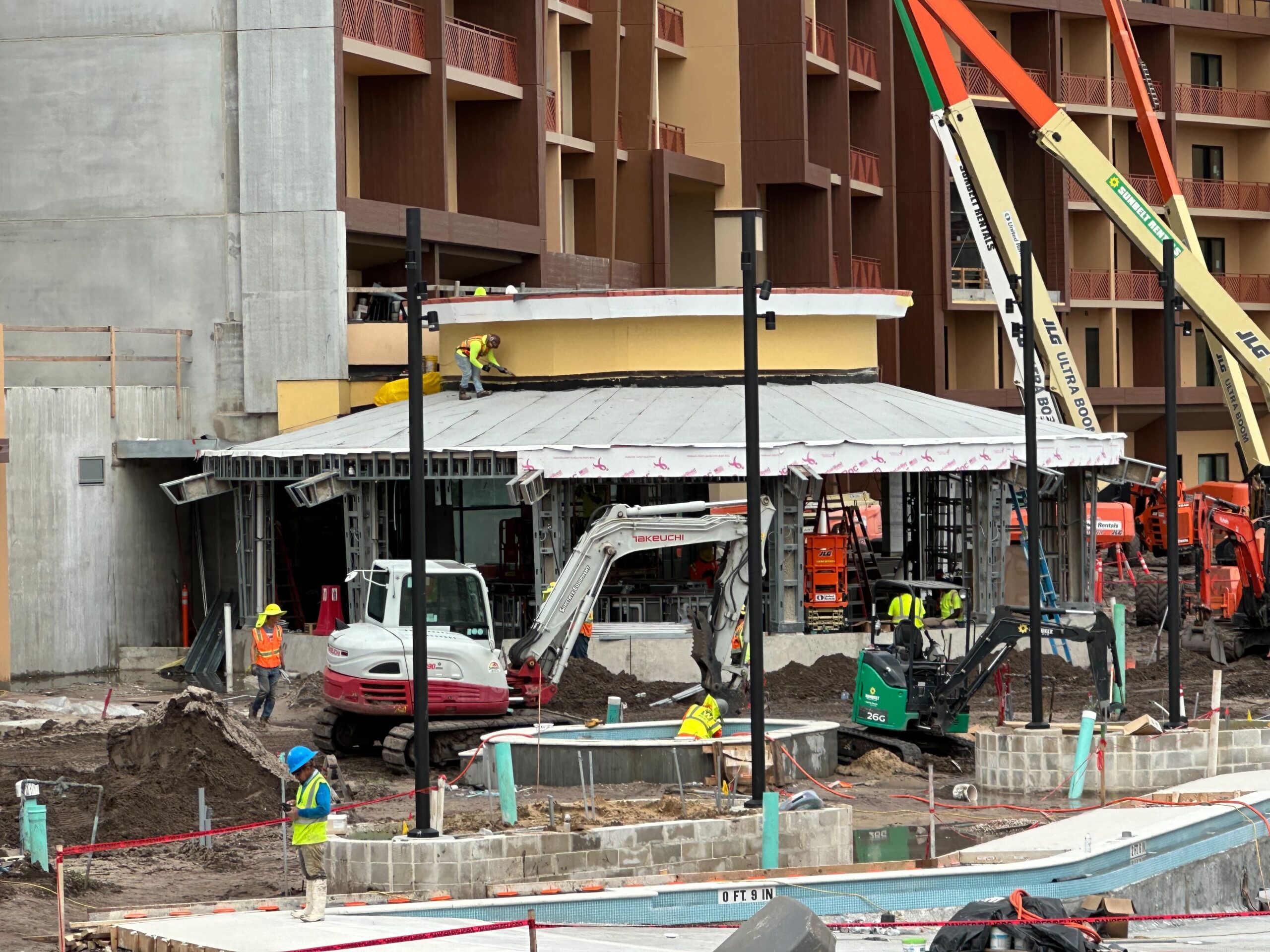 Construction workers and machinery are active at a building site with a partially constructed structure and several cranes and excavators in use.