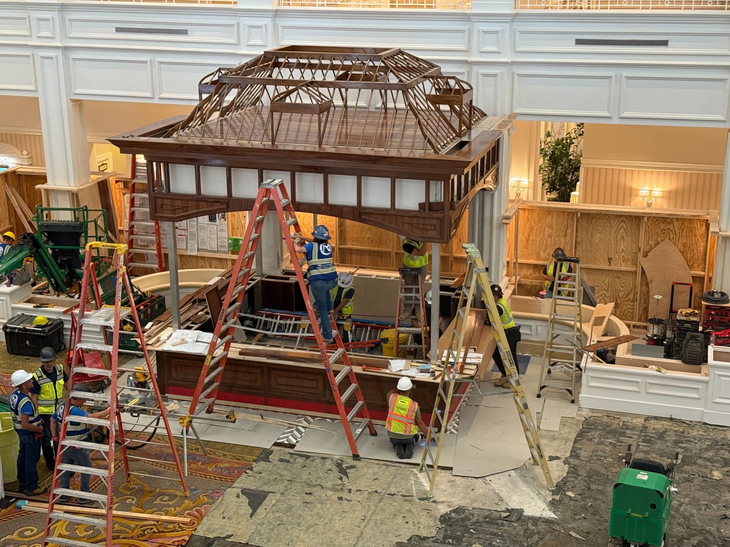 Workers in safety gear construct a large wooden gazebo-like structure indoors, surrounded by ladders and building materials.