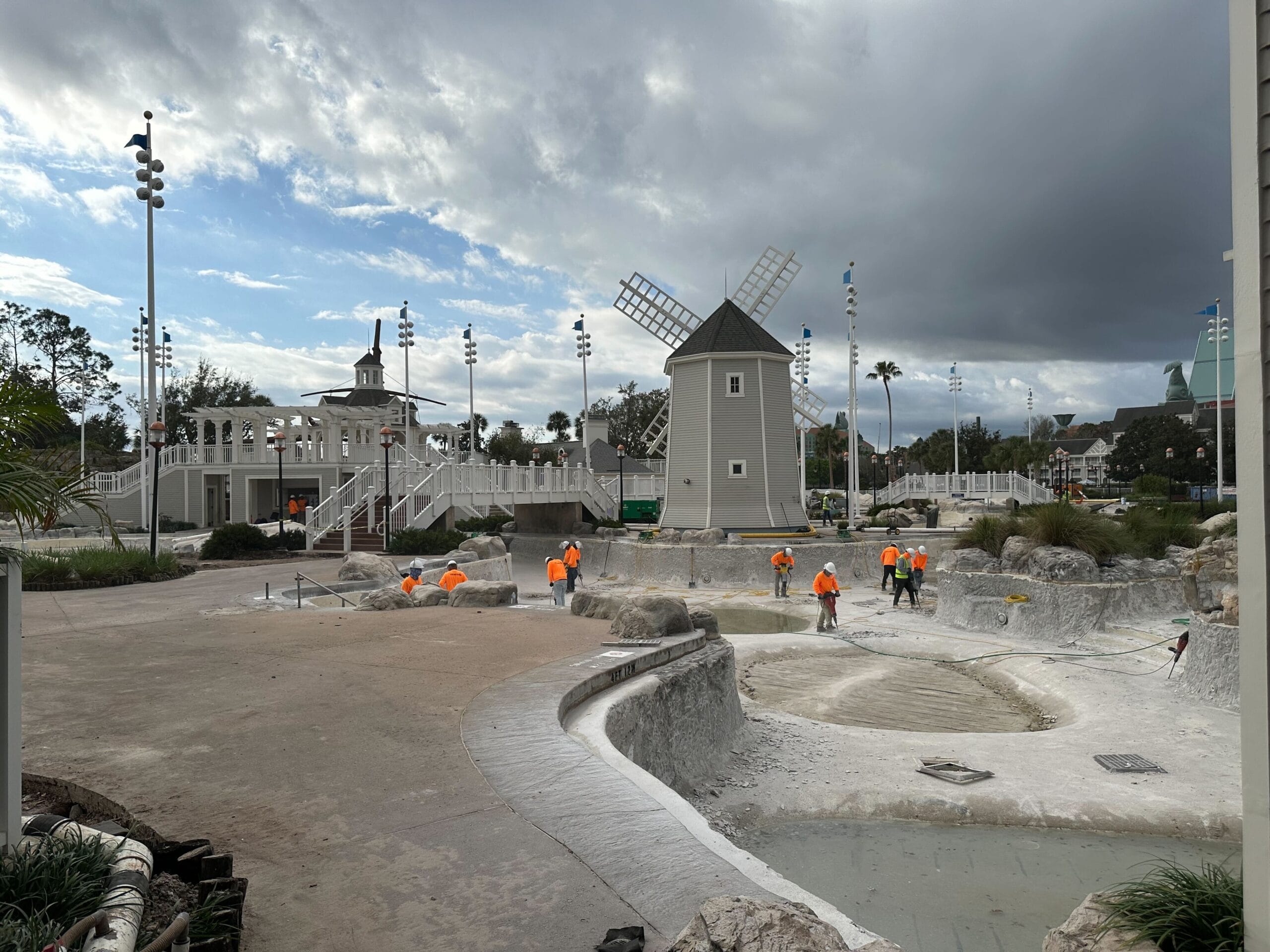 Workers in orange vests perform maintenance on an empty swimming pool near a windmill structure, under a cloudy sky.