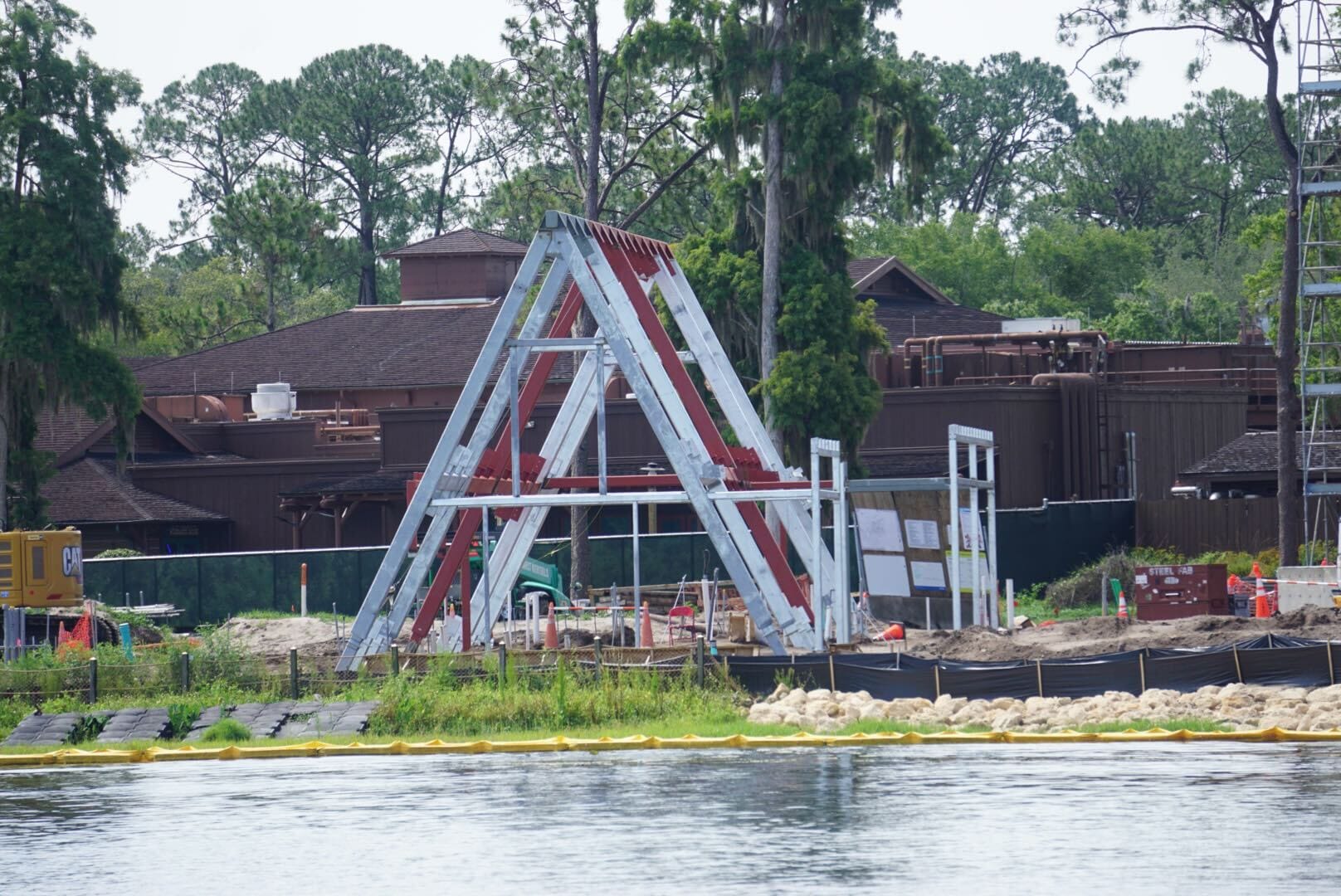 A metal A-frame structure under construction near water, with trees and brown buildings in the background.