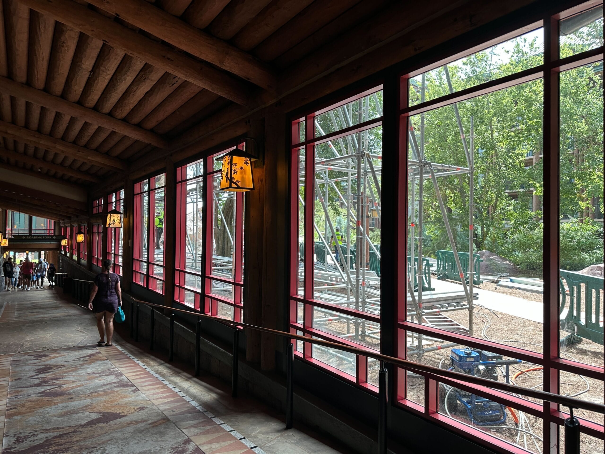 A covered walkway with large red-framed windows, looking out to trees and construction scaffolding outside.
