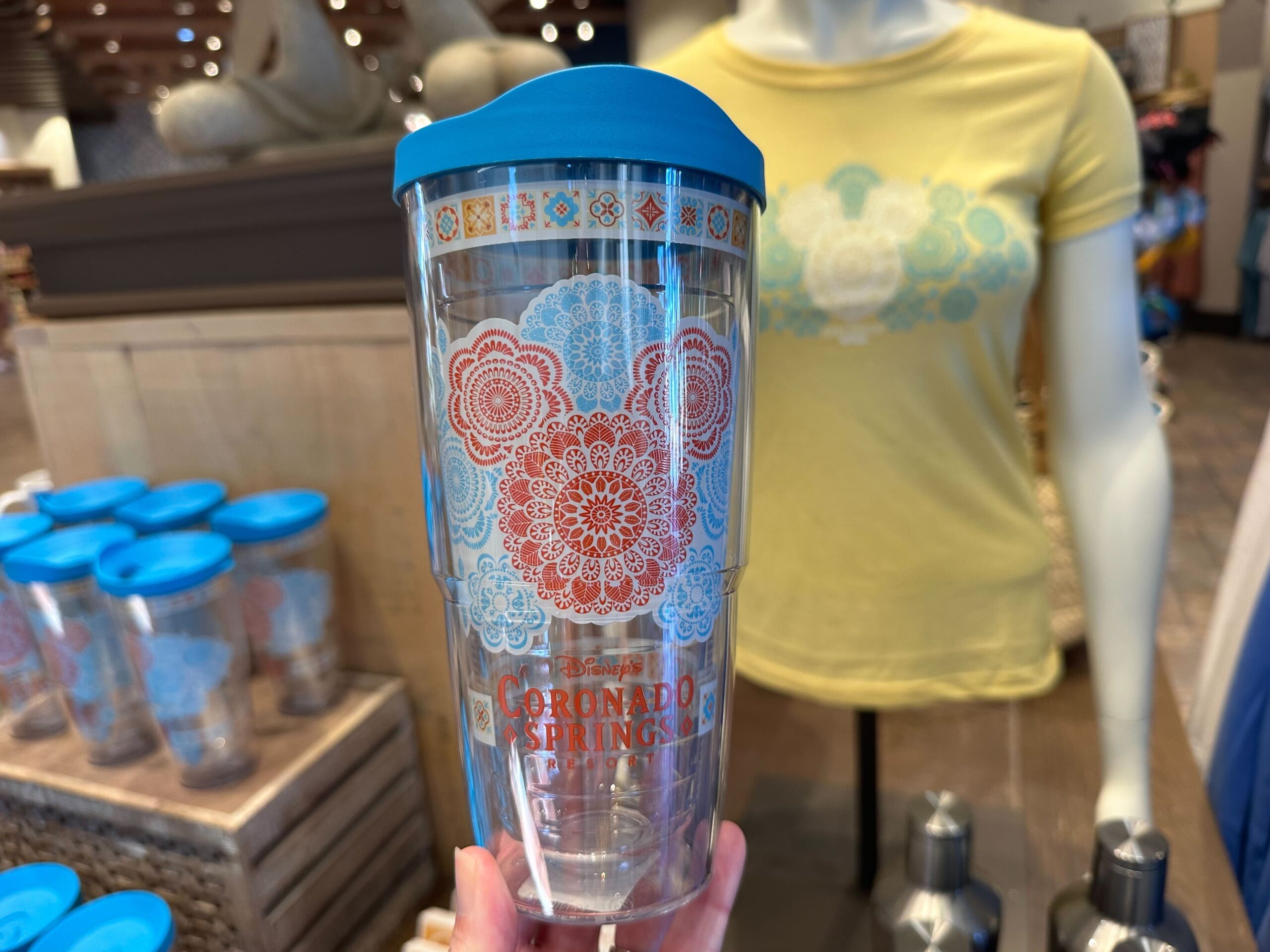 Clear travel mug with blue lid, featuring intricate floral designs and Coronado Springs Resort text. Displayed among Coronado Springs merchandise, it sits next to a matching T-shirt, creating the perfect pairing for fans.