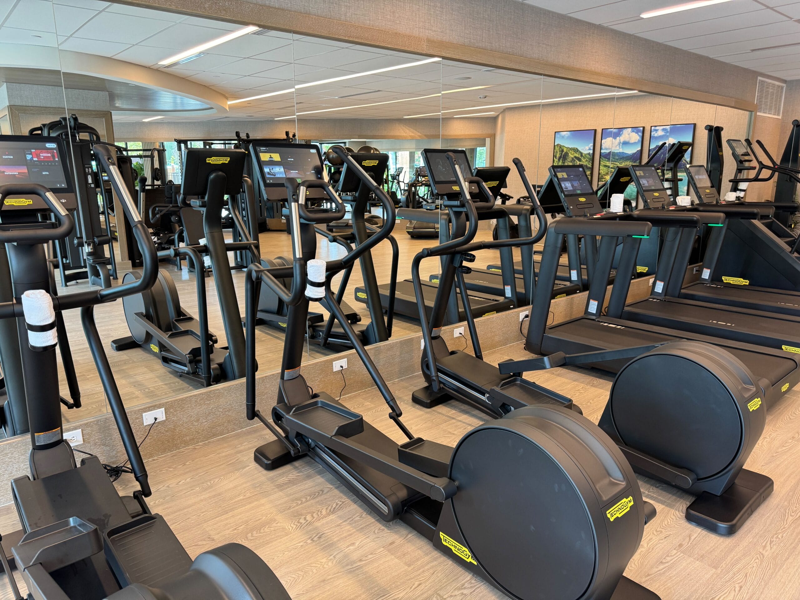 A modern gym with several elliptical machines in front of a mirrored wall, surrounded by wooden flooring and large windows.