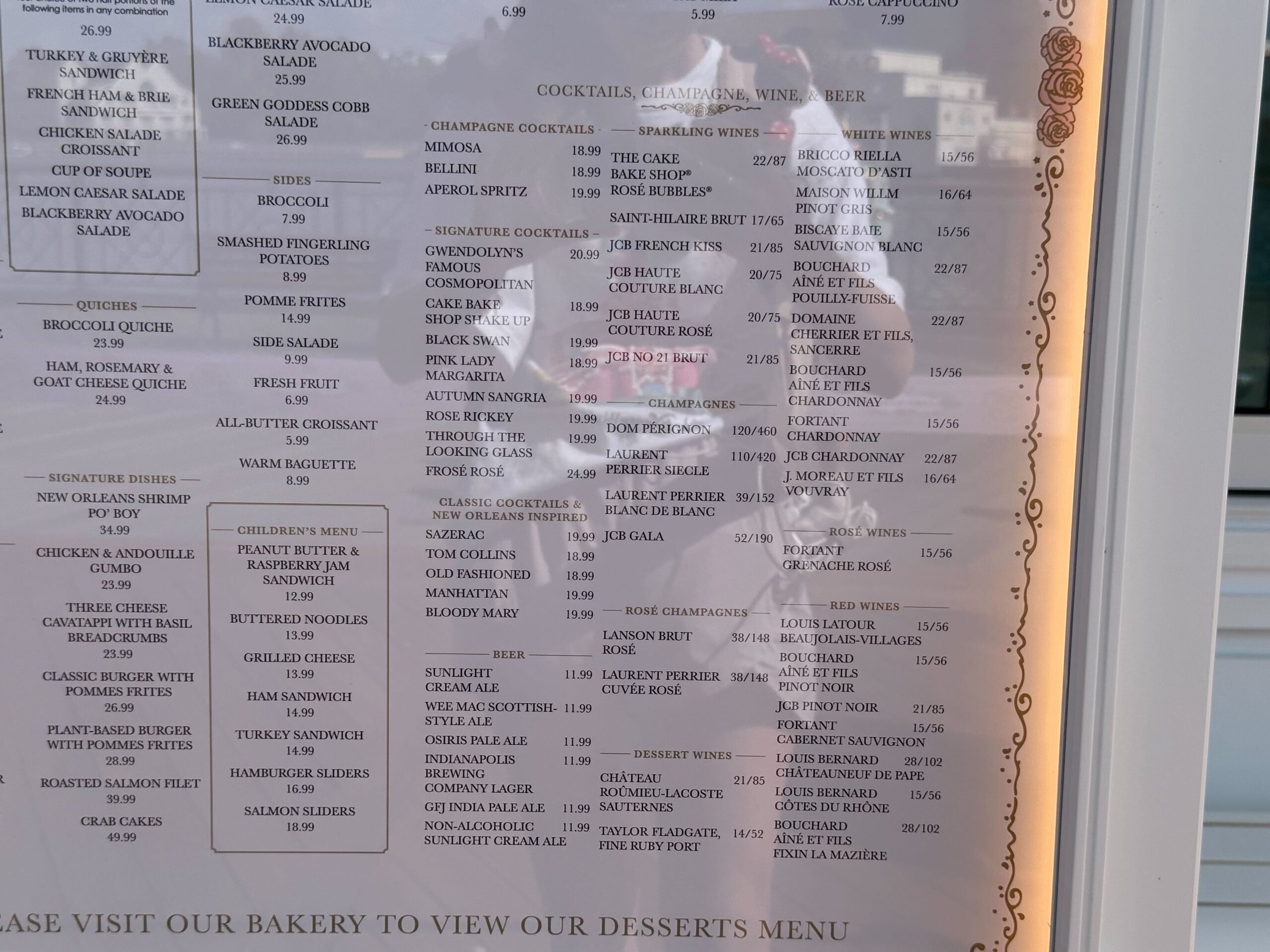 A restaurant menu, reminiscent of a cake bake shop's charm, is displayed on the window, listing salads, soups, cocktails, wine, and beer with prices. Reflections of people mingle with the enticing offerings on the glass.