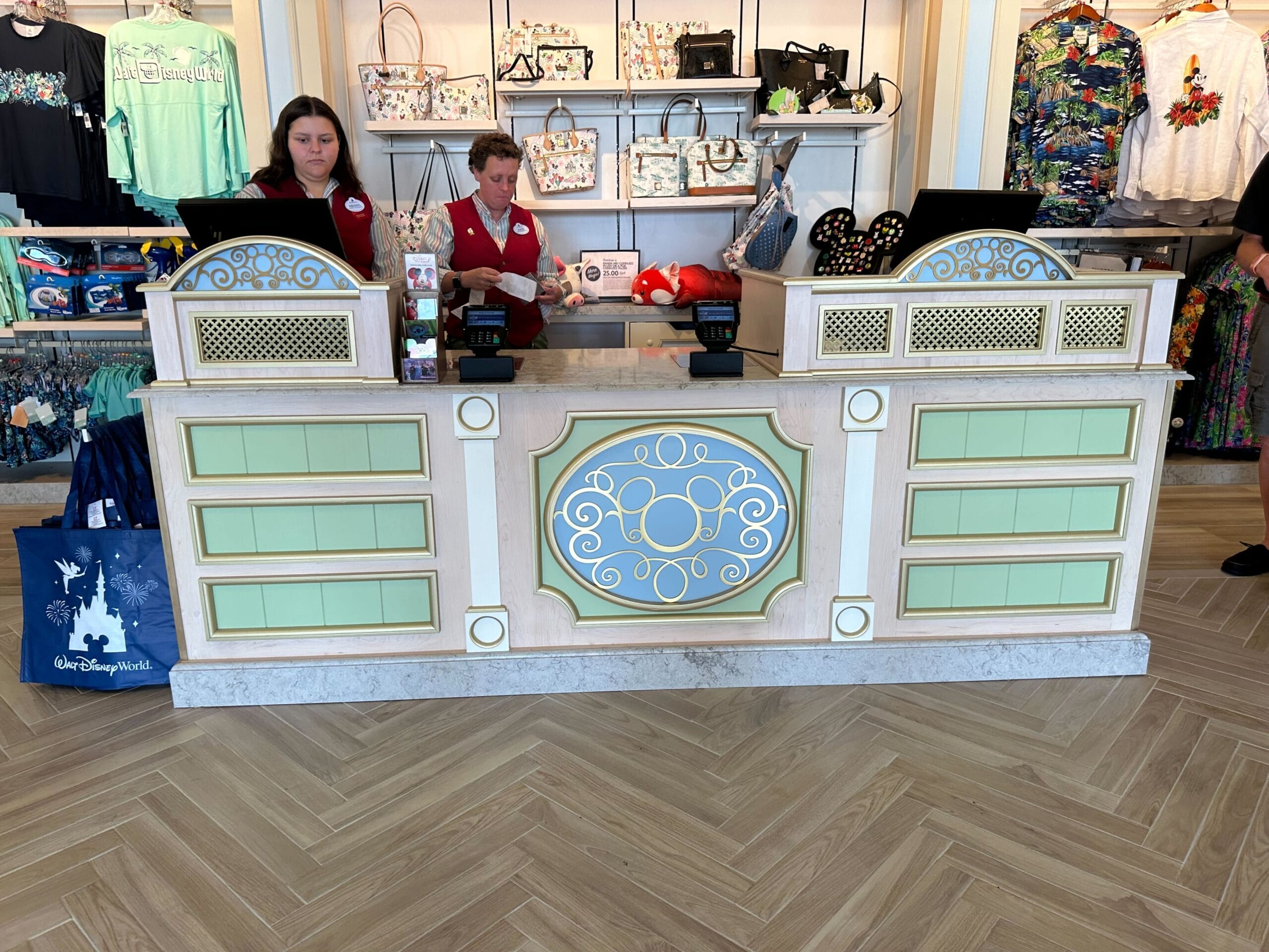 Two employees stand behind a decorative store counter at Disney's Boardwalk Inn Resort, surrounded by merchandise like bags and shirts. Signs and cash registers are neatly arranged, echoing the charm of the Screen Door shop atmosphere.