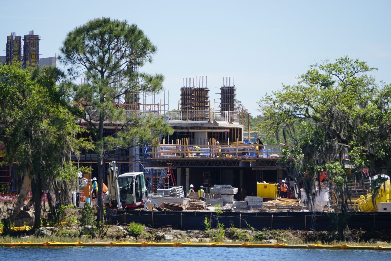 Construction site by a river with workers, machinery, and building materials. Partially built structure in the center, surrounded by trees and a clear sky.