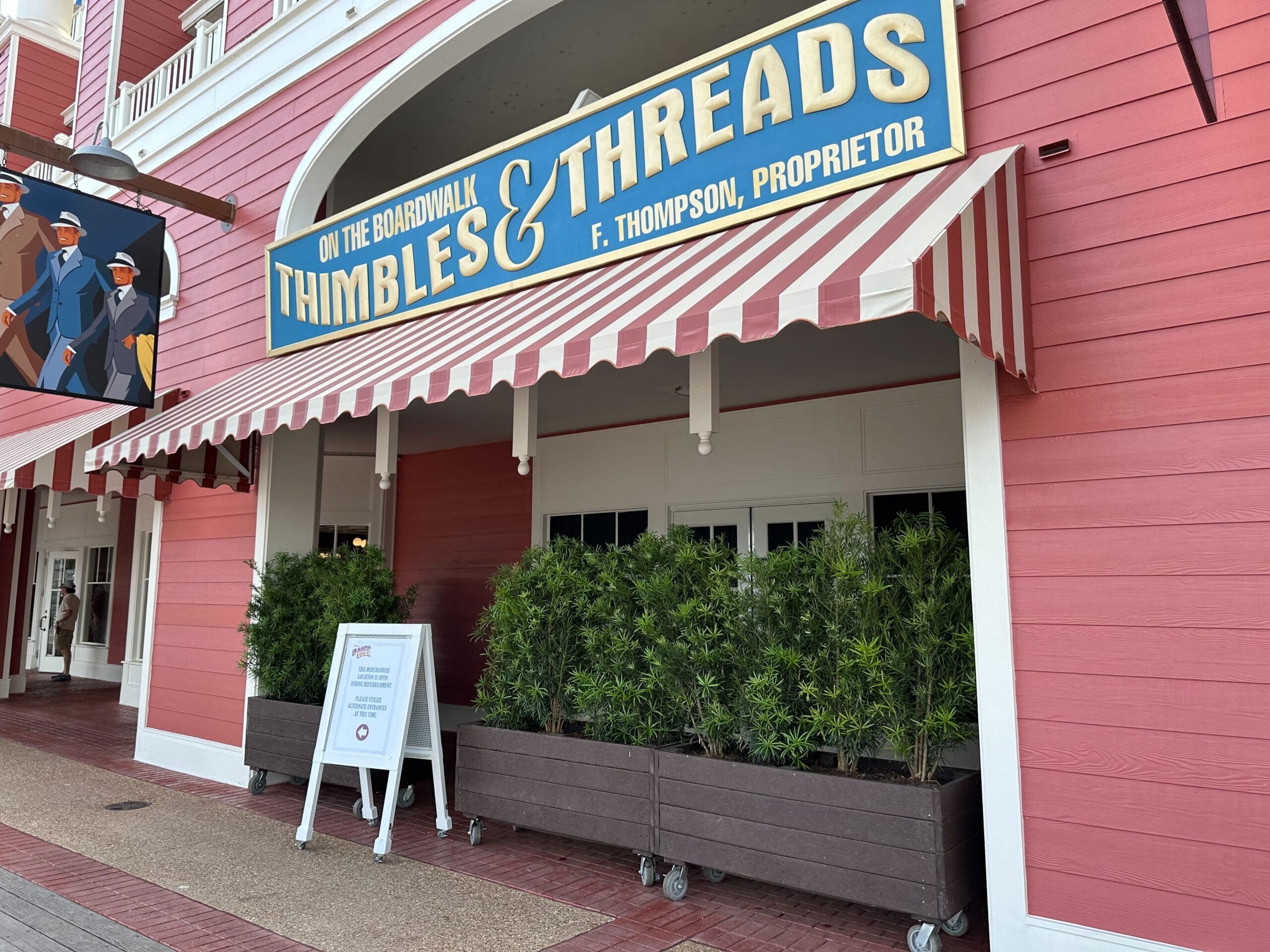 The storefront of Thimbles & Threads at Disney's Boardwalk Inn Resort boasts a striped awning, pink exterior, greenery in planters, and a charming signboard, reminiscent of the quaint allure found in the Screen Door General Store's renovated section.