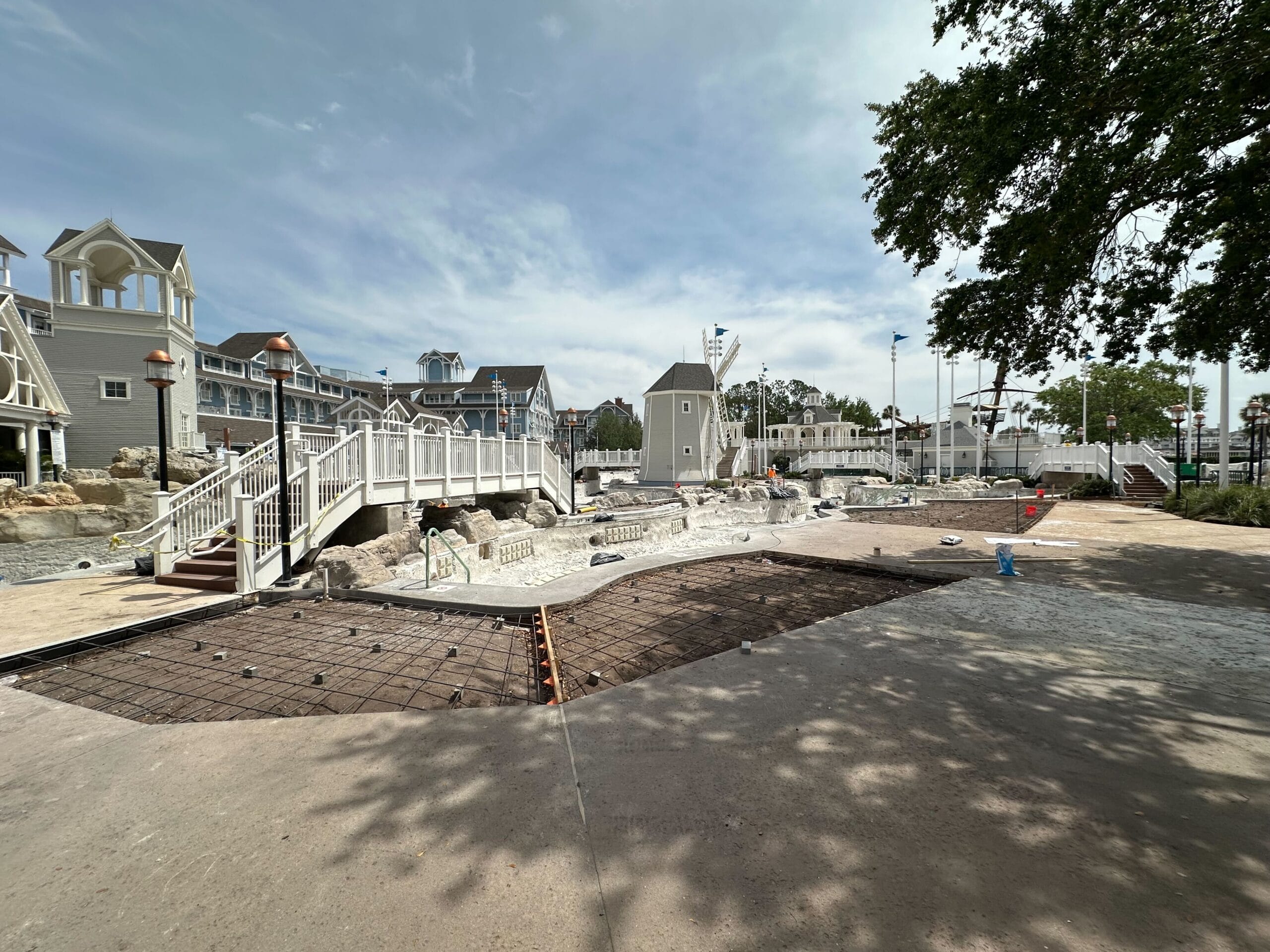 Outdoor construction site with partially completed concrete work, near a series of large buildings with white railings at Disney's Yacht Club. It is a sunny day, and there are trees overhead, as scaffolding rises to shape the future structure.