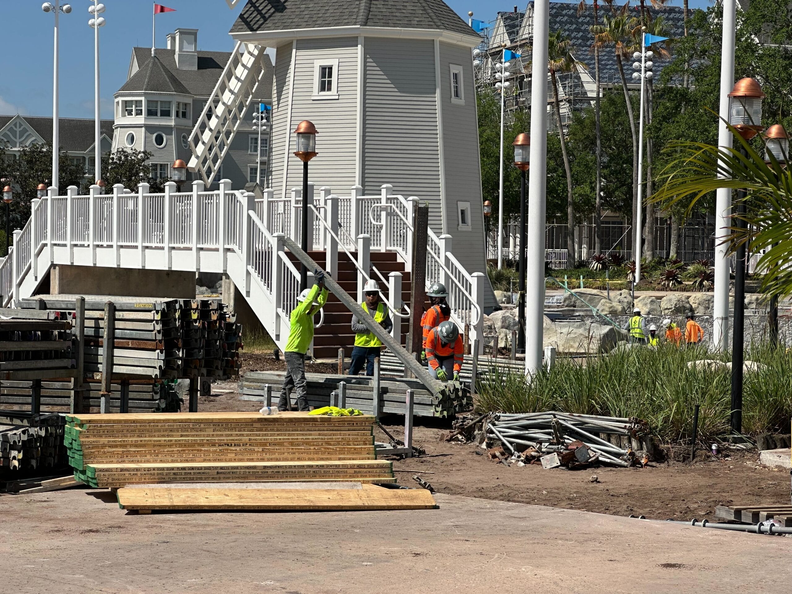 Workers in safety gear are busy at the construction site near a wooden structure and piles of materials, while a tower of new scaffolding rises impressively. Behind them, a white building stands proudly amid carefully designed landscaping.