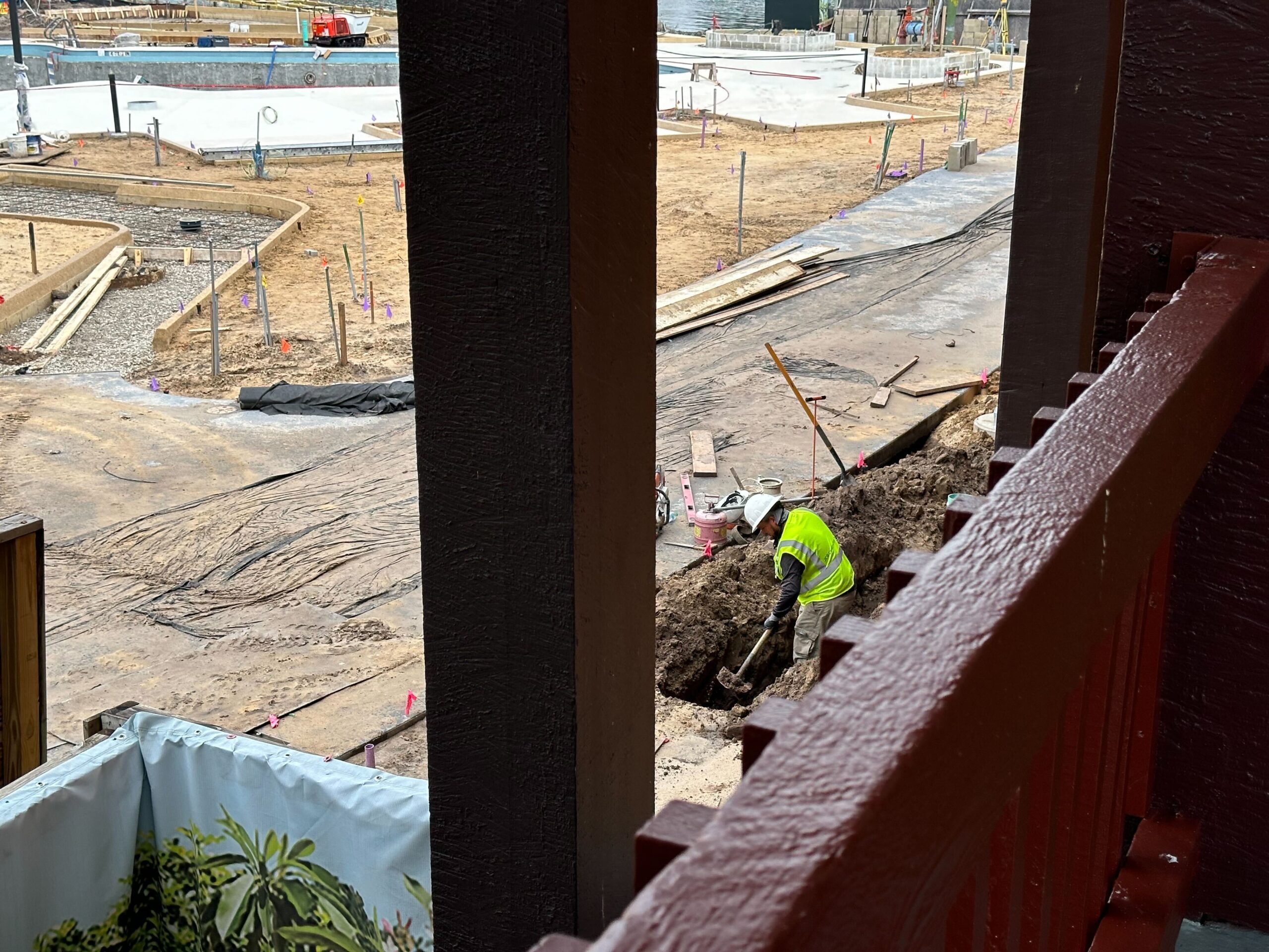 Construction worker wearing a reflective vest digging a trench in a large construction site viewed from a nearby structure with a railing in the foreground.