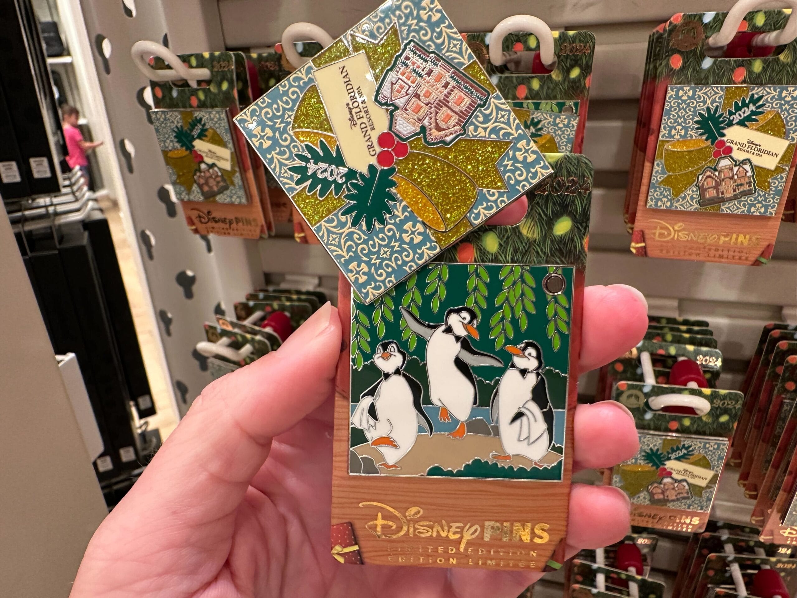 Hand holding a Disney pin featuring penguins under mistletoe, with a display of similar pins in the background.