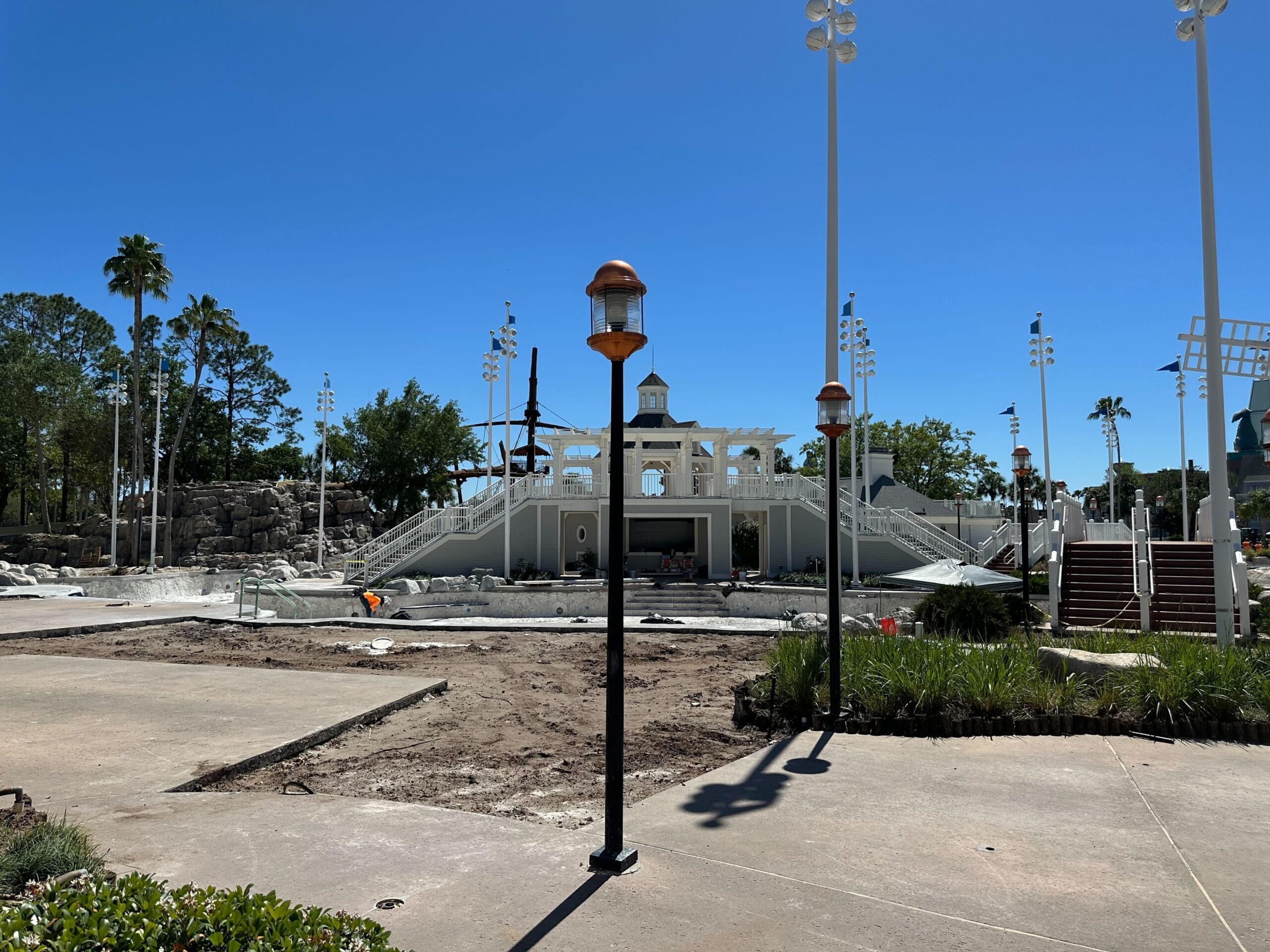 View of a construction site near Disney’s Yacht Club, featuring a partially built structure surrounded by lights and trees. A clear blue sky is in the background, and the ground appears mostly dirt with patches of grass—much like the serene surroundings of Stormalong Bay Pool.