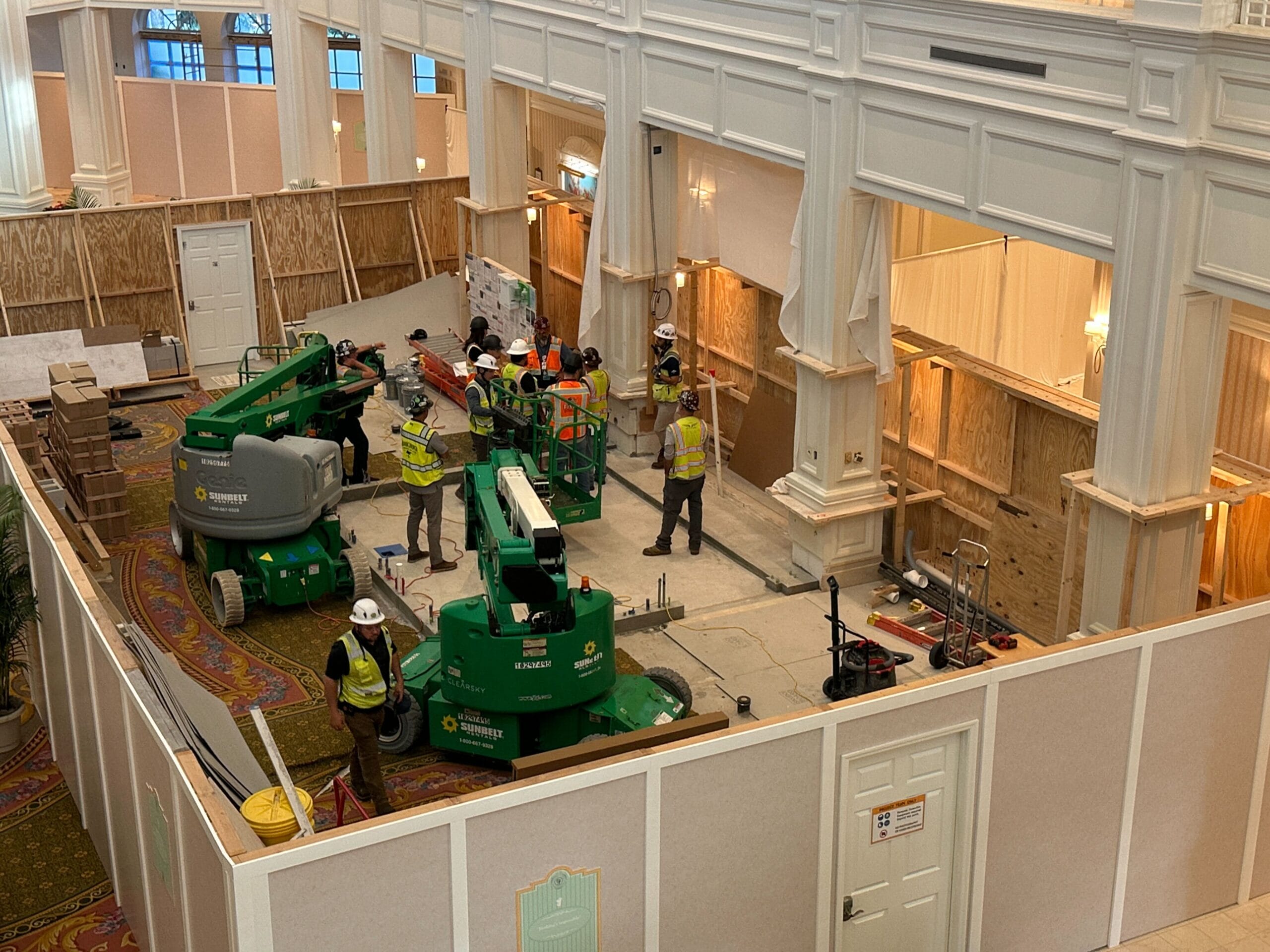 Construction workers in safety gear operate machinery inside a sectioned-off area of an ornate building interior.